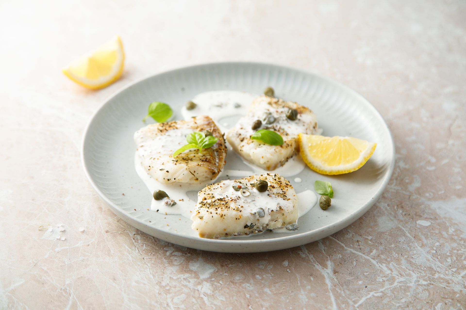 White fish fillets garnished with herbs on a plate, with lemon wedges on a light tabletop.