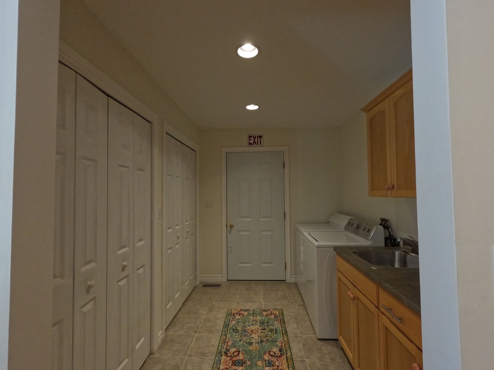 Narrow laundry room with white closet doors, wood cabinets, washer, sink, and patterned rug.