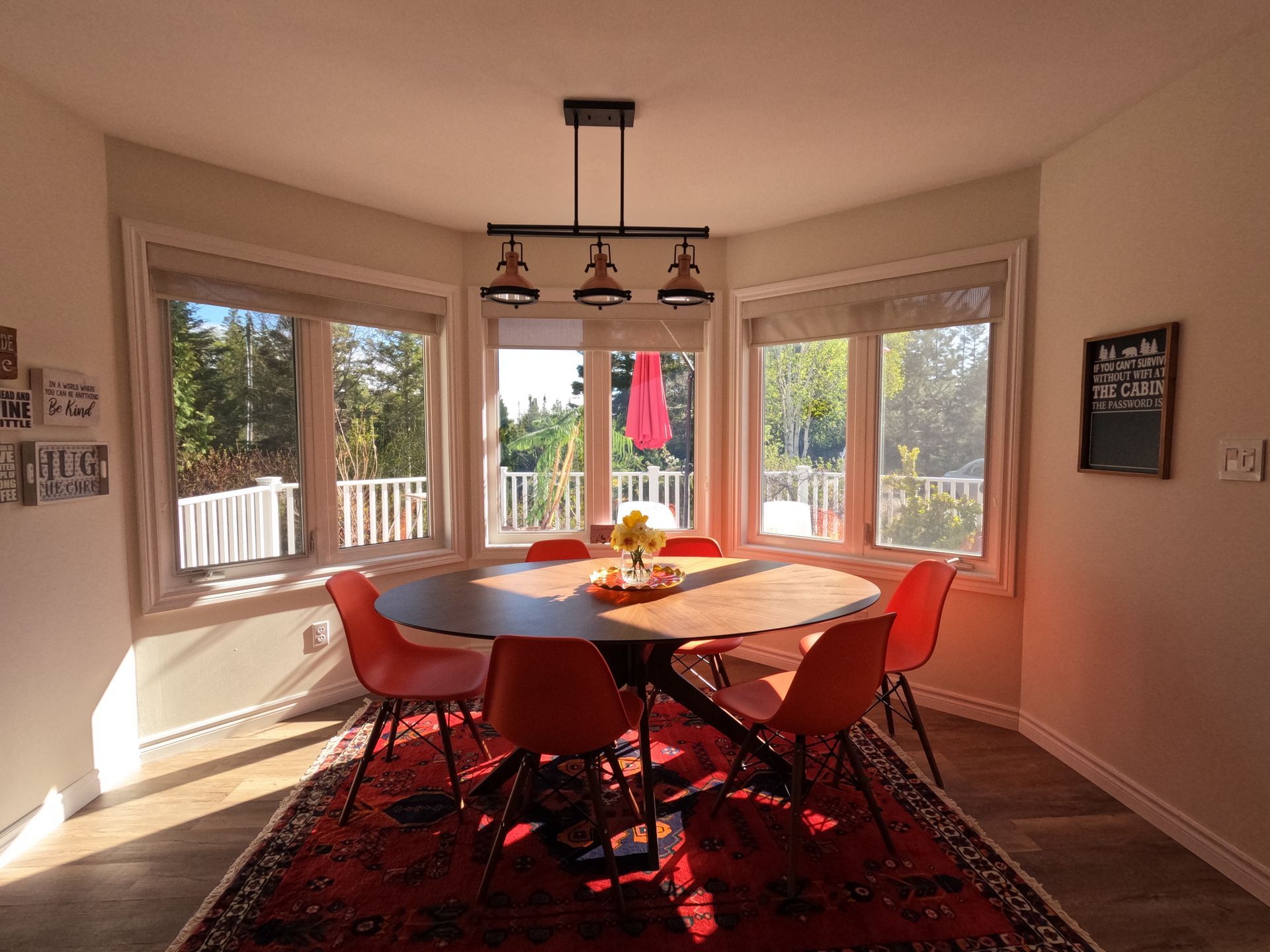Bright dining room with red chairs, wooden table, bay windows, and sunlight on a patterned rug.