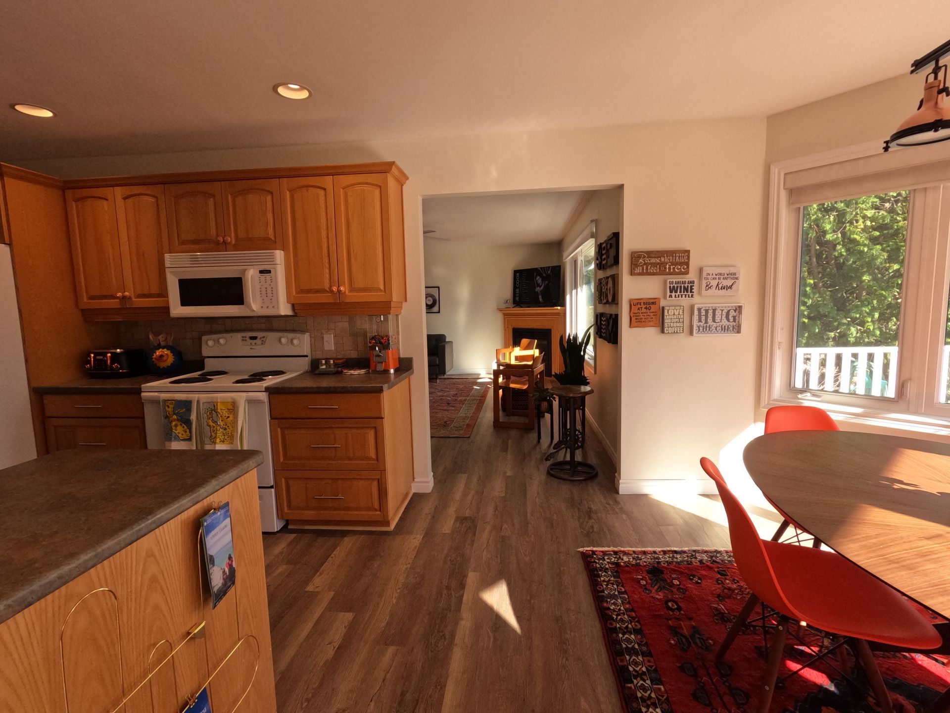 Bright kitchen with wood cabinets, white appliances, and sunlight streaming into a dining area with orange chairs.