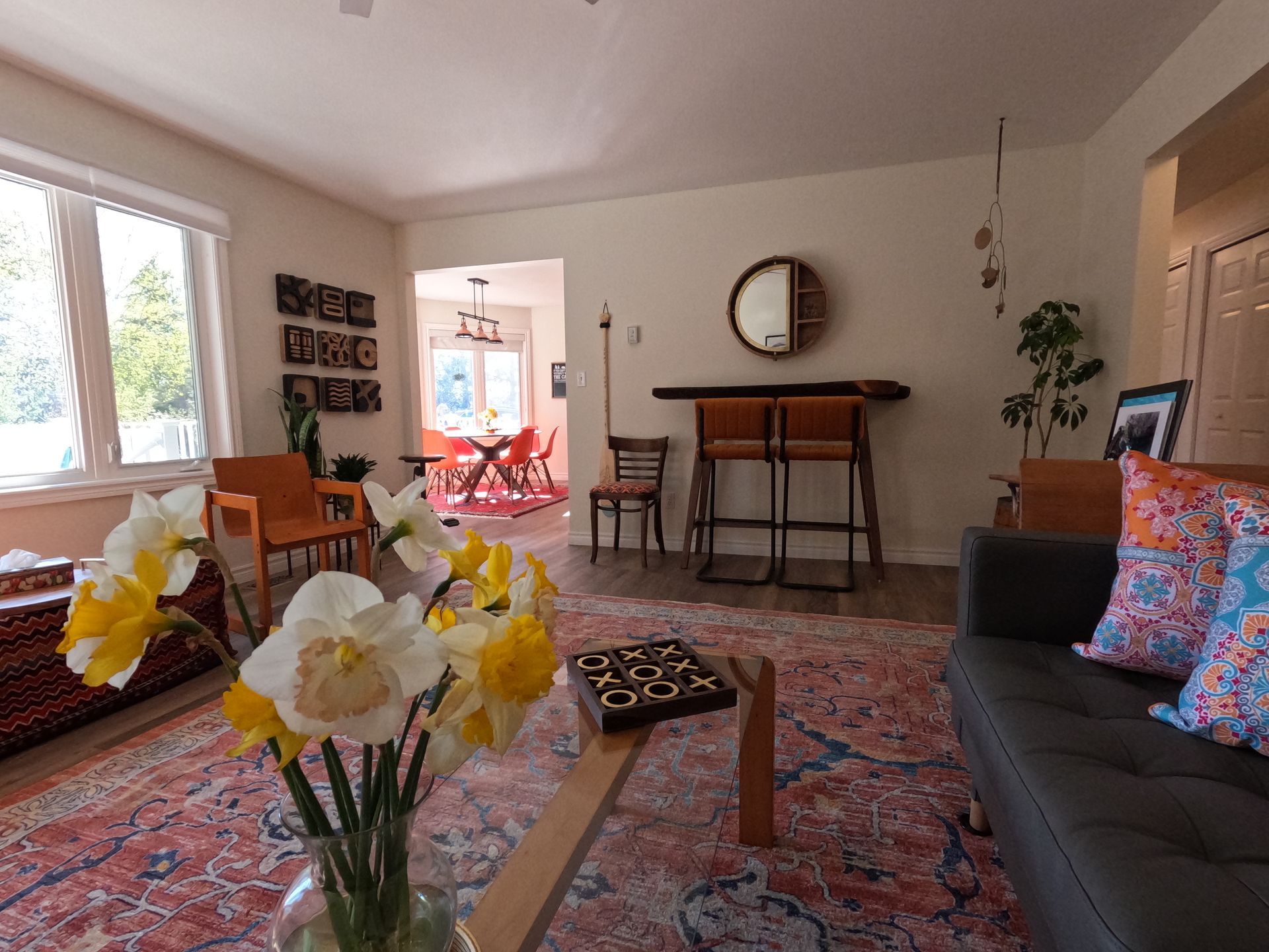 Bright living room with patterned rug, gray sofa, flowers, dining table, and a large wall clock.