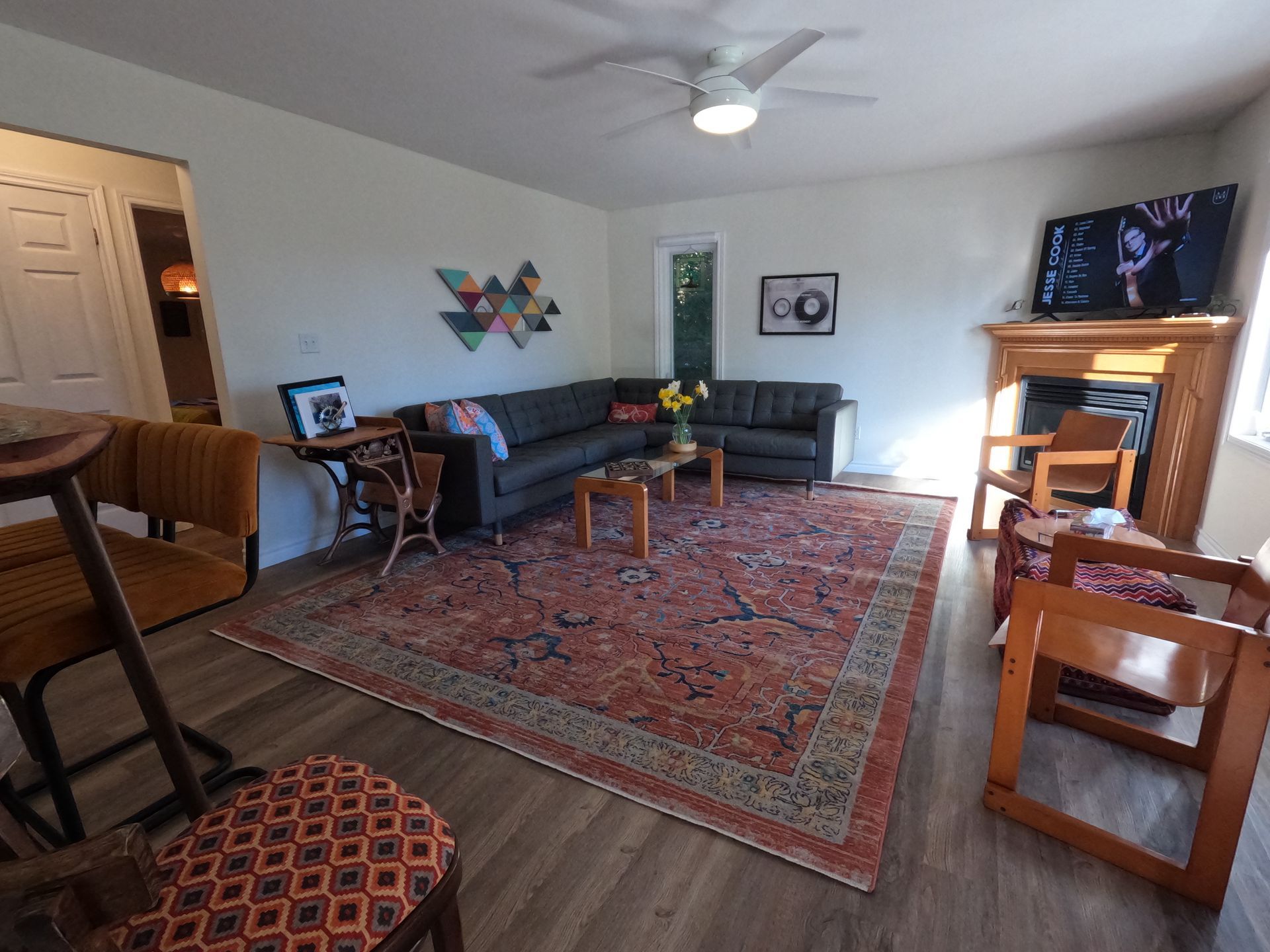 Living room with sofas, patterned rug, fireplace, and TV on a mantel, lit by a ceiling fan.