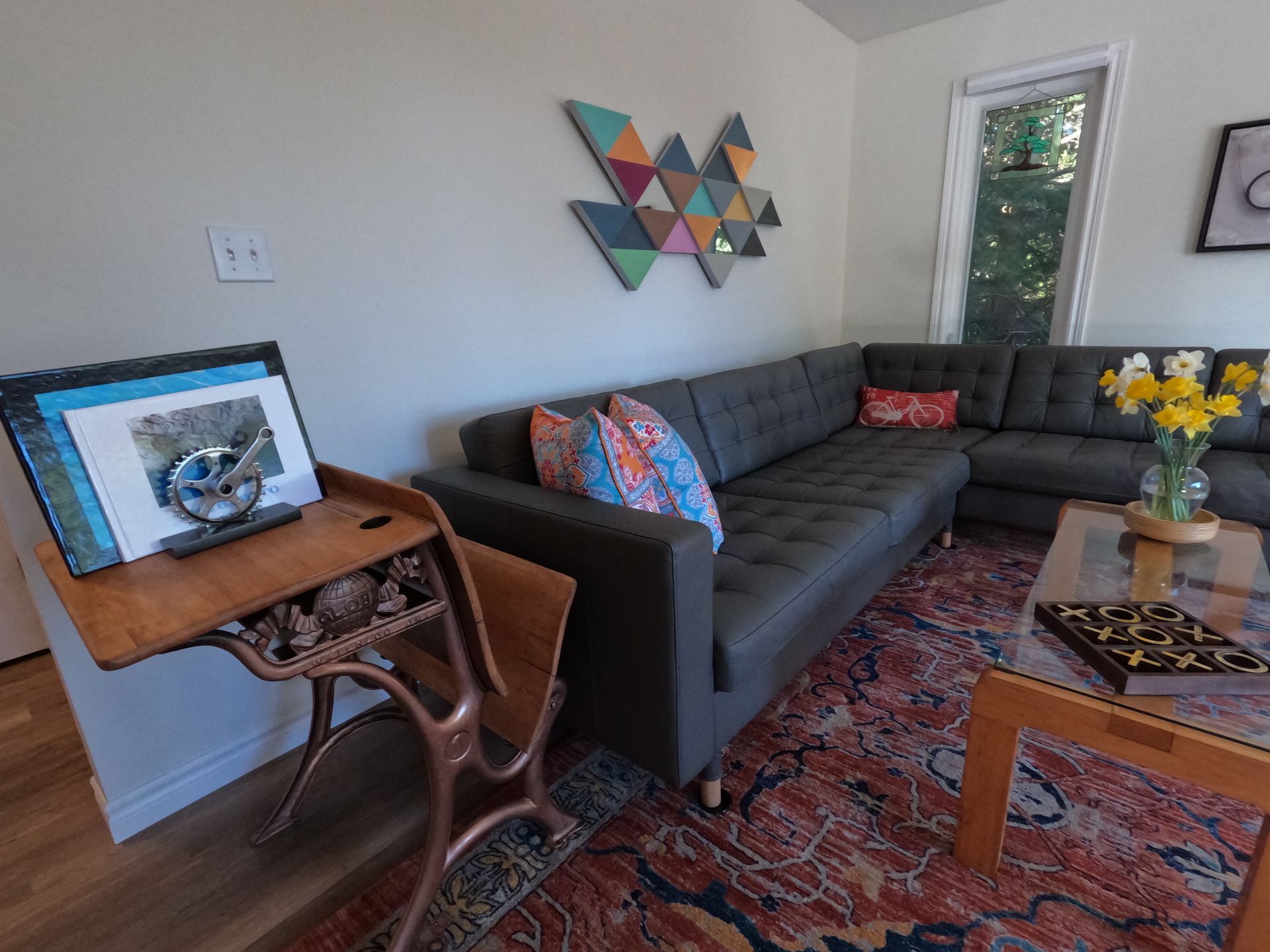Living room with gray sectional, patterned rug, side table, wall art, and a laptop on a wooden desk