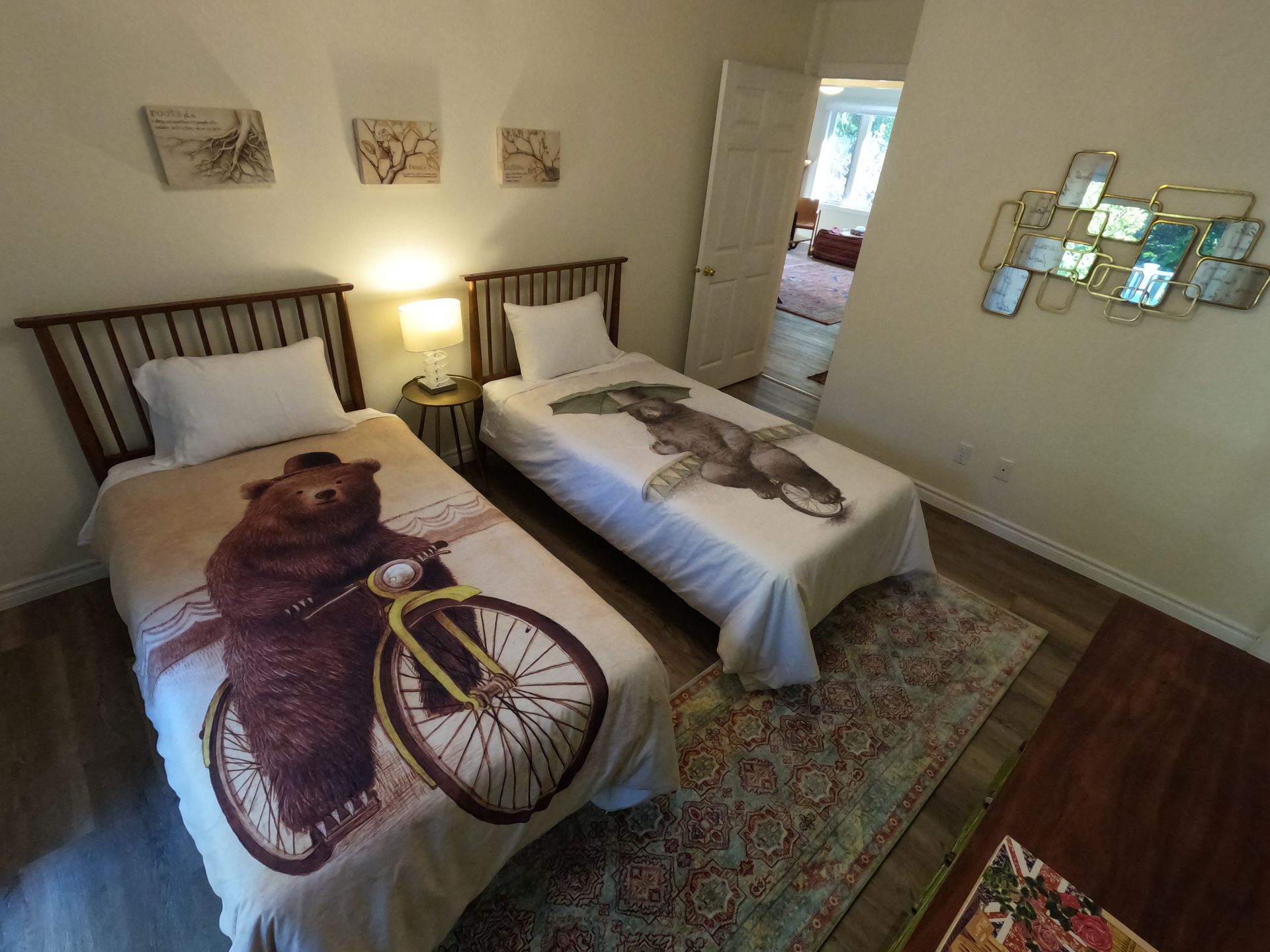 Two twin beds in a beige bedroom, one with a bear and bicycle blanket, bedside lamp, patterned rug.