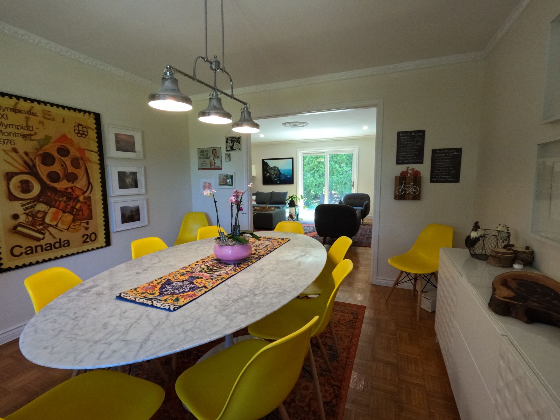 Bright dining room with a long table, yellow chairs, hanging lights, and wall art overlooking a garden.
