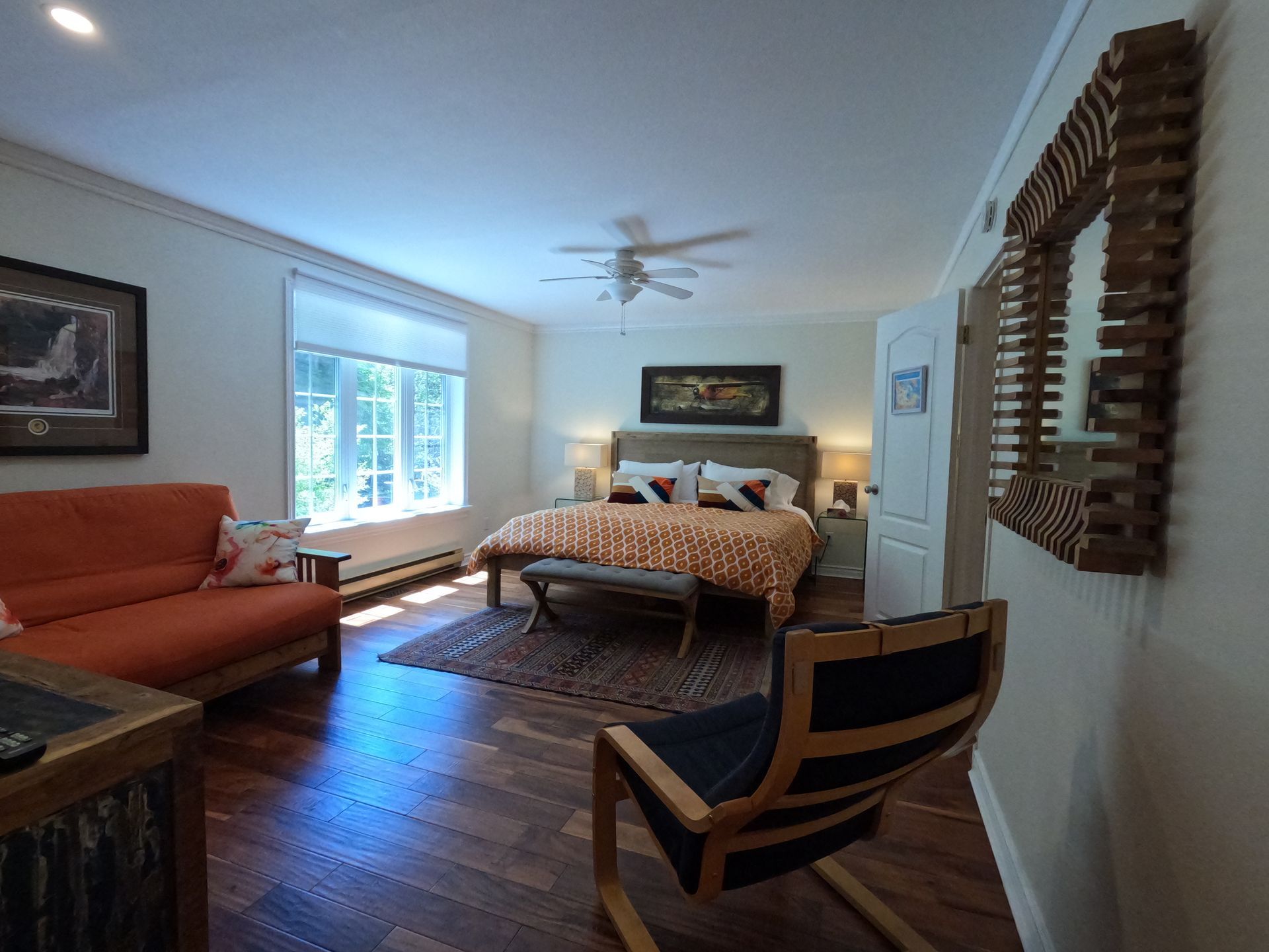 Bright bedroom with a bed, orange sofa, wooden floor, window light, and a ceiling fan