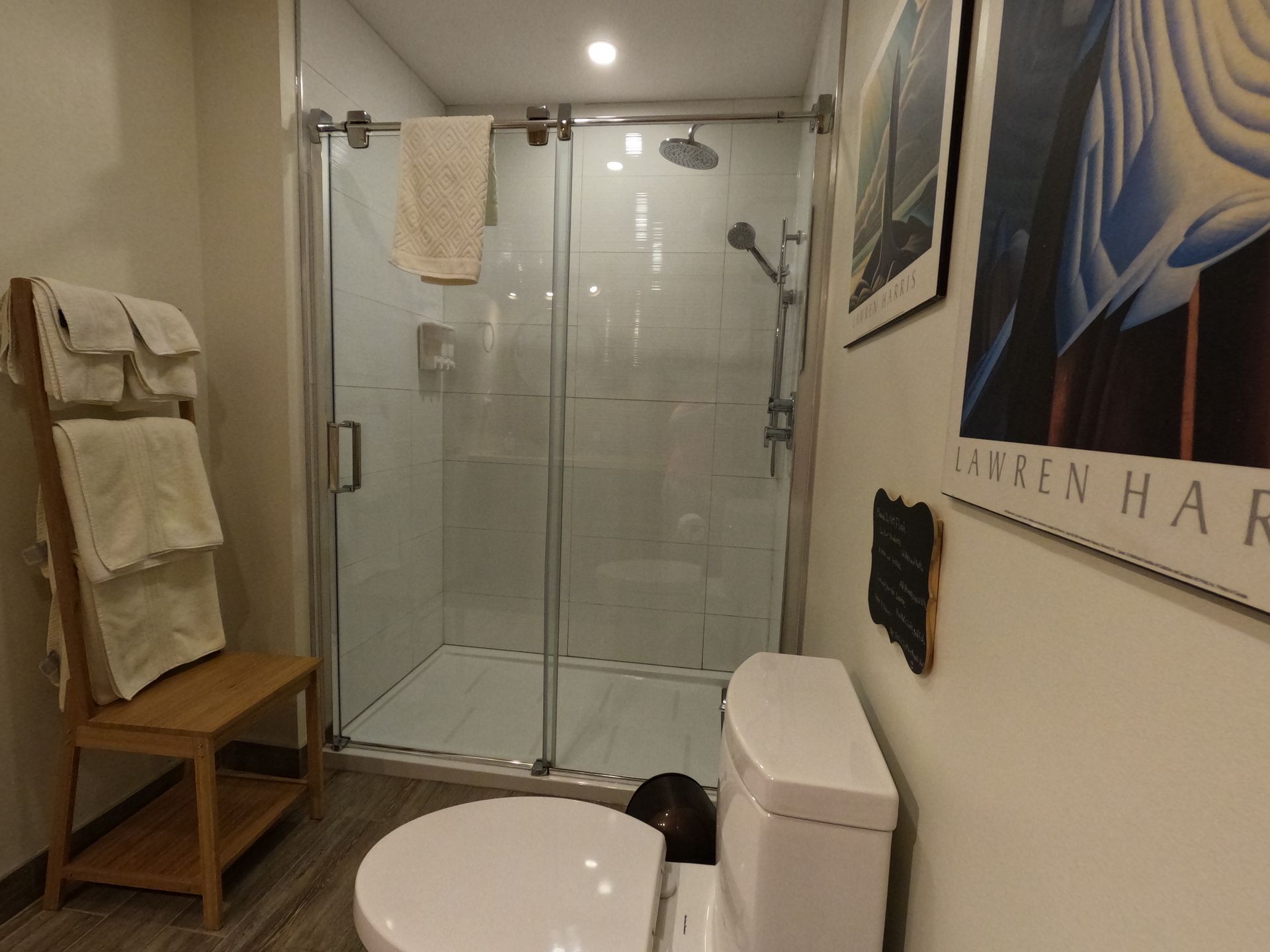 Small bathroom with glass shower, white toilet, round rug, towels, and wall art.
