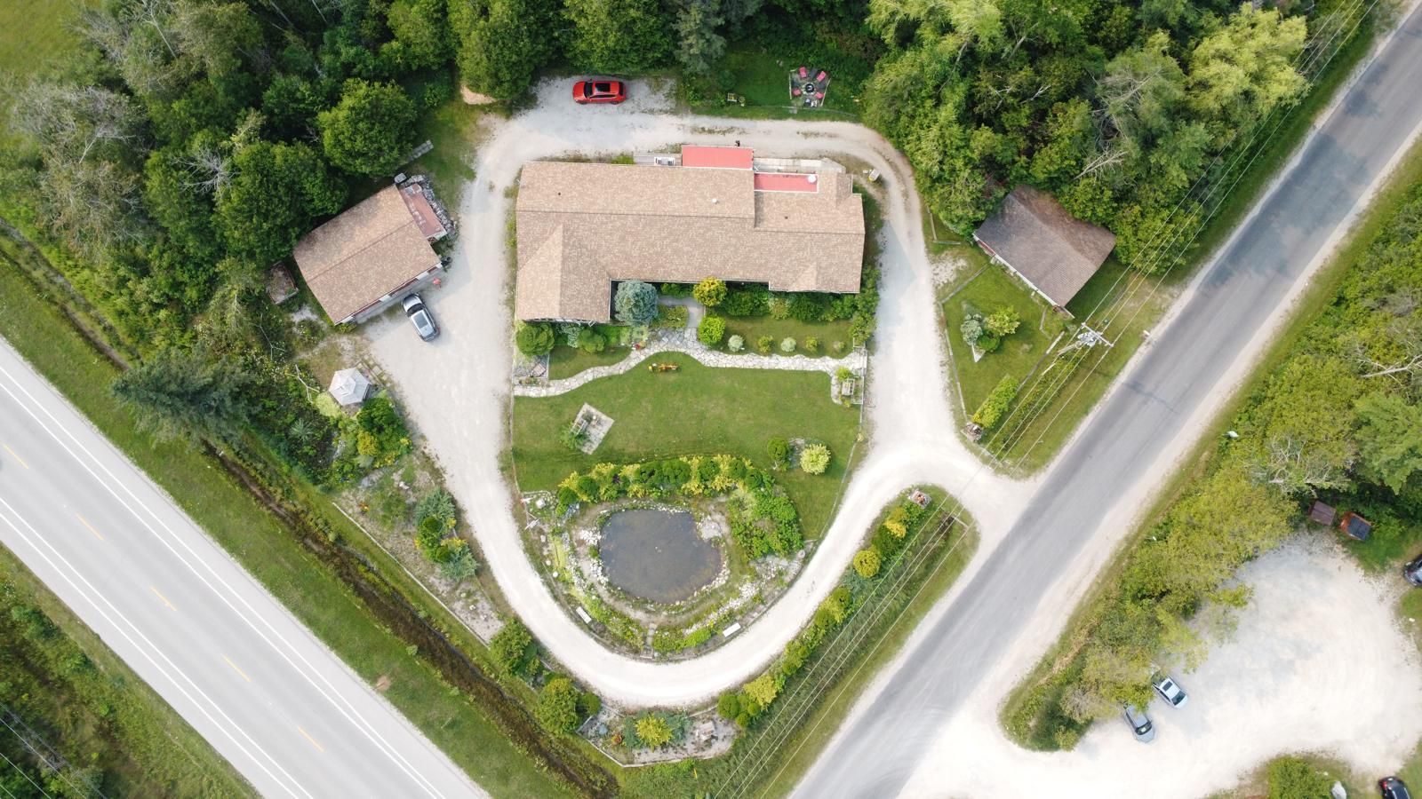 Aerial view of a small house and yard at a road intersection, surrounded by trees and gravel driveways