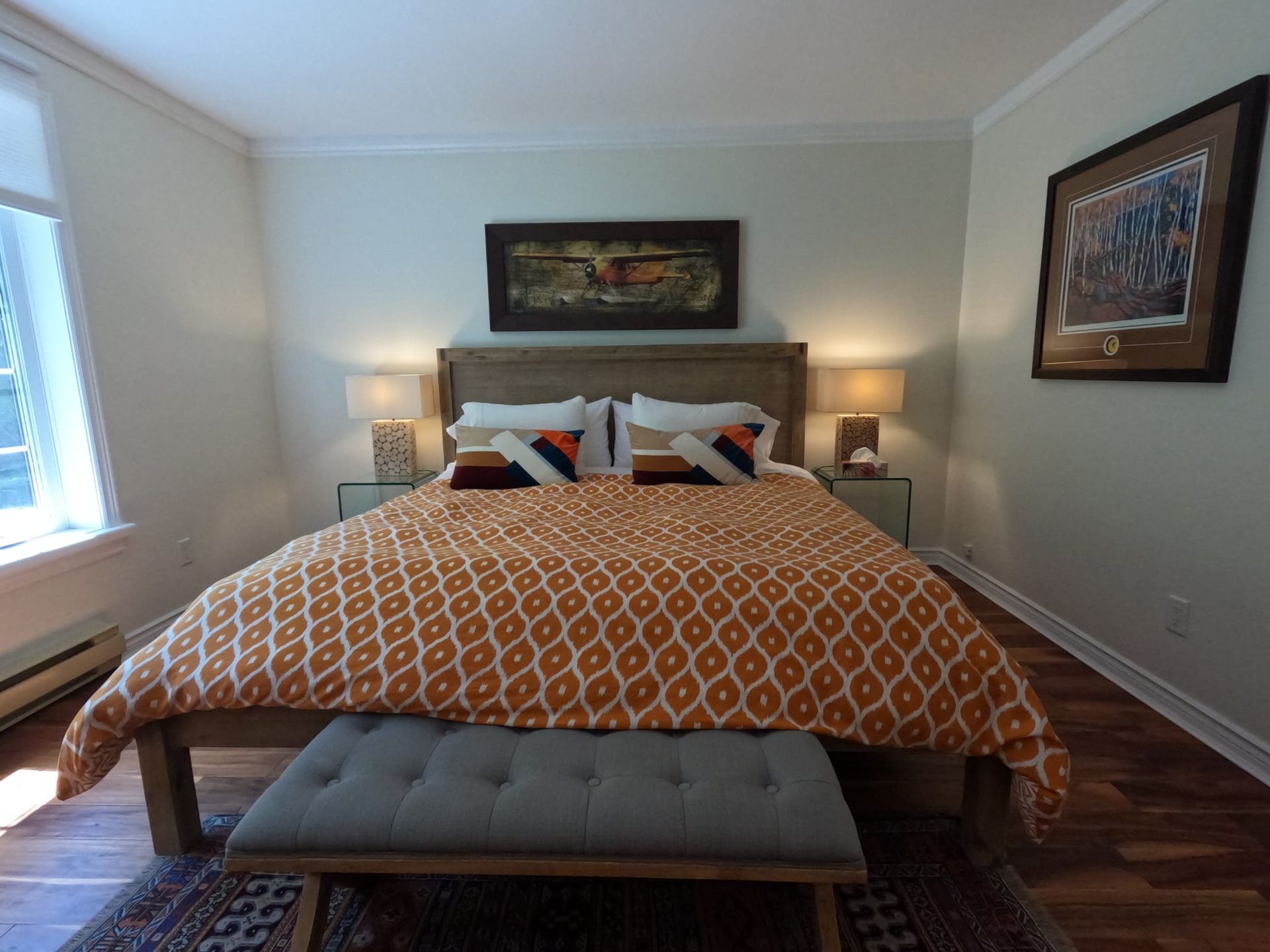 Cozy bedroom with a made bed, orange patterned duvet, bedside lamps, and framed wall art.