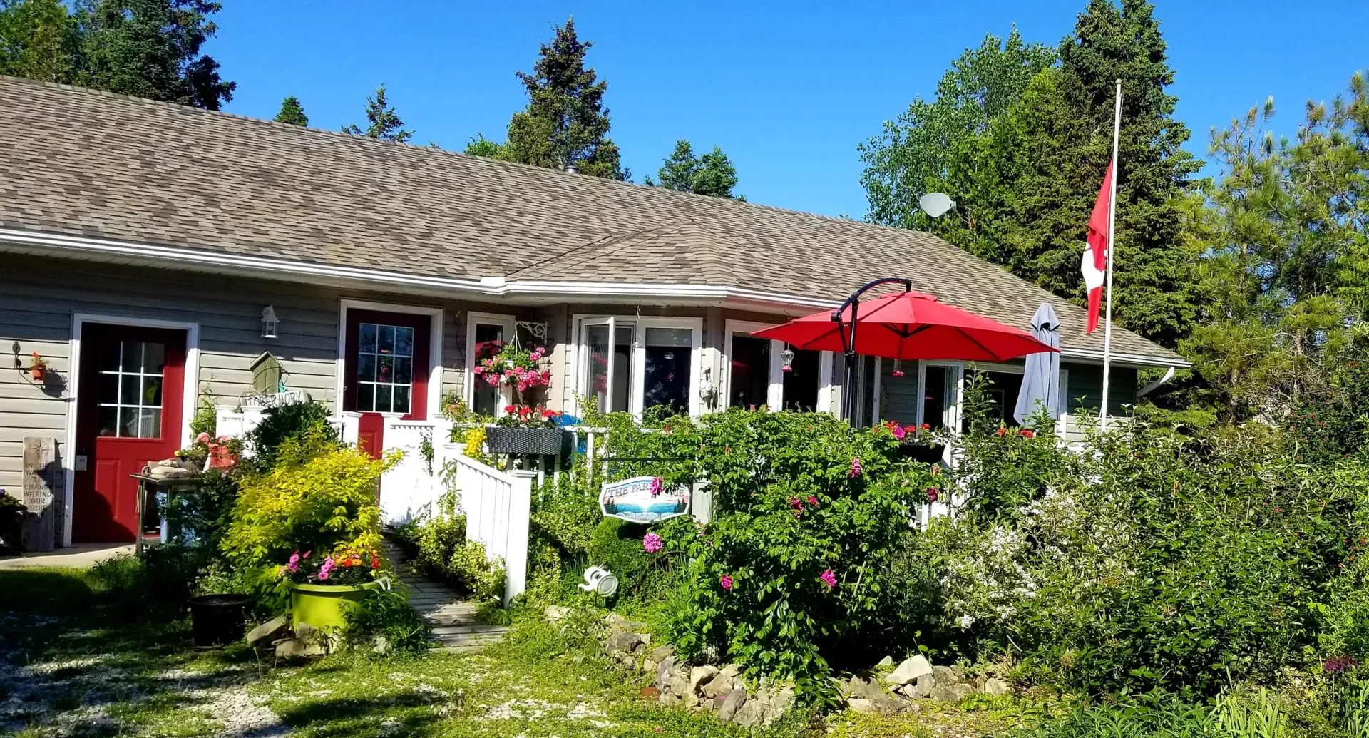 Sunlit garden with a rustic house, red patio umbrella, and Canadian flag under a blue sky