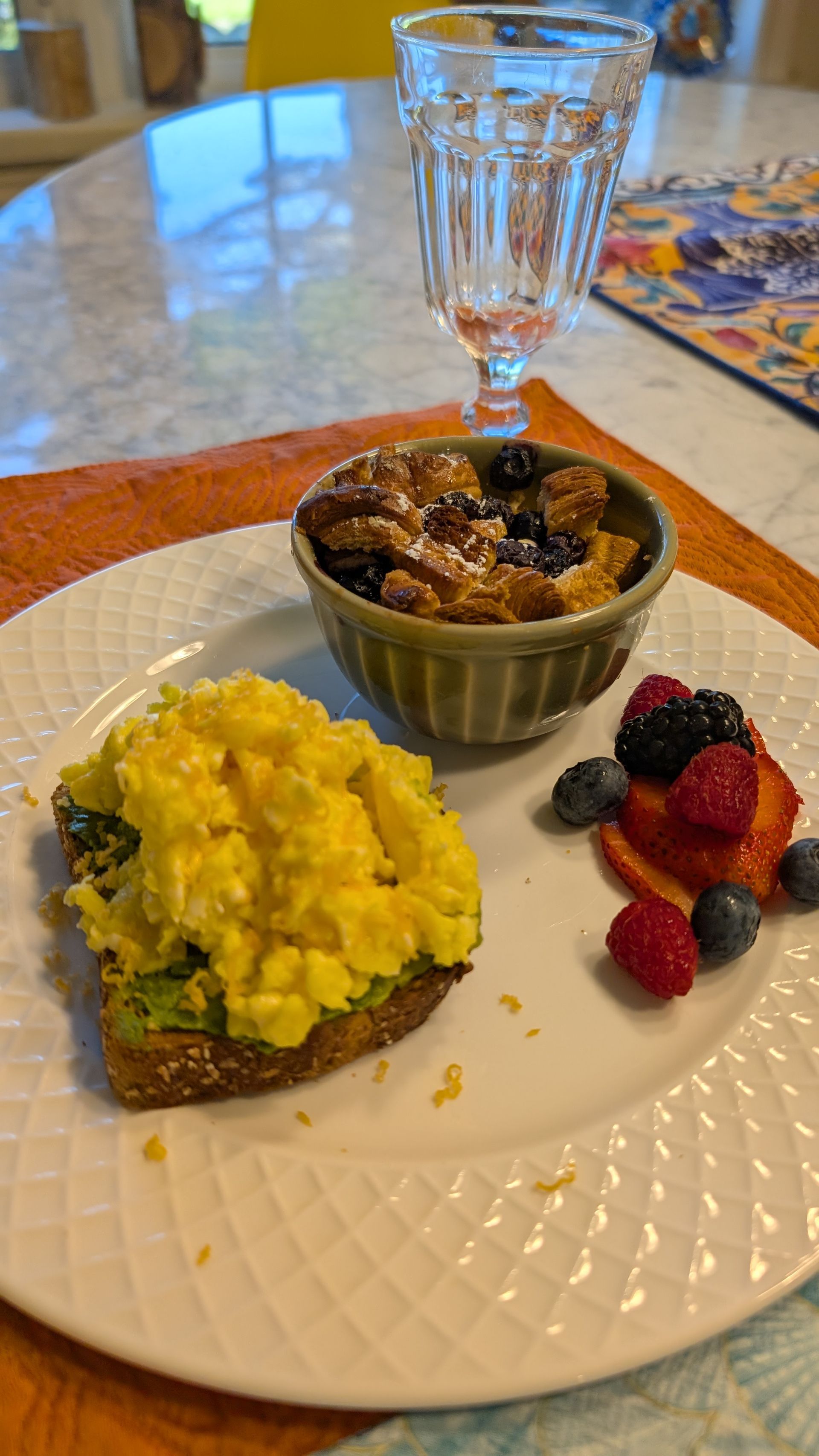 Plated breakfast with scrambled eggs, toast, fruit, and a small baked dish on a table with a water glass.