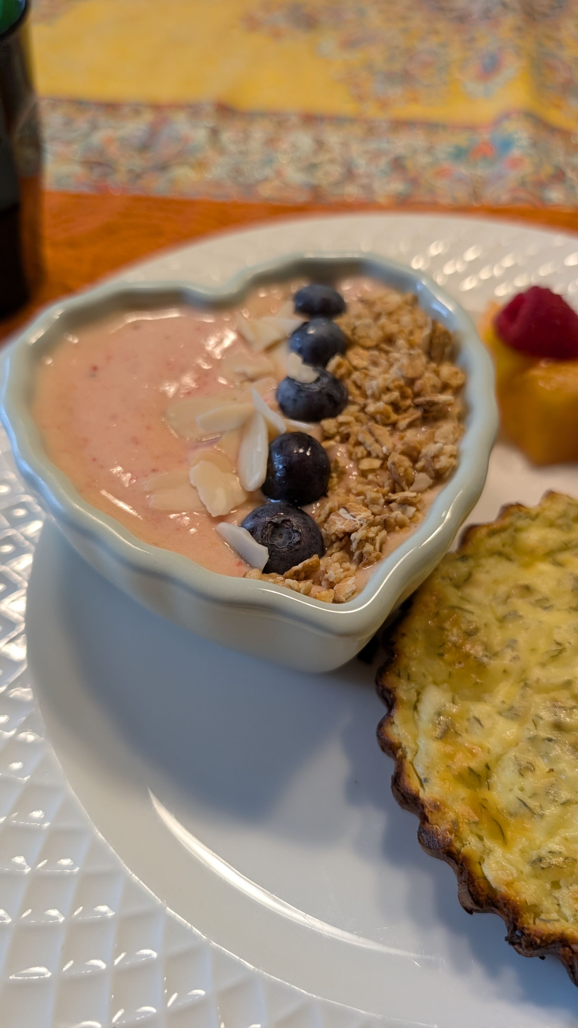 Heart-shaped breakfast bowl with pink yogurt, blueberries, granola, and two pastries on a white plate