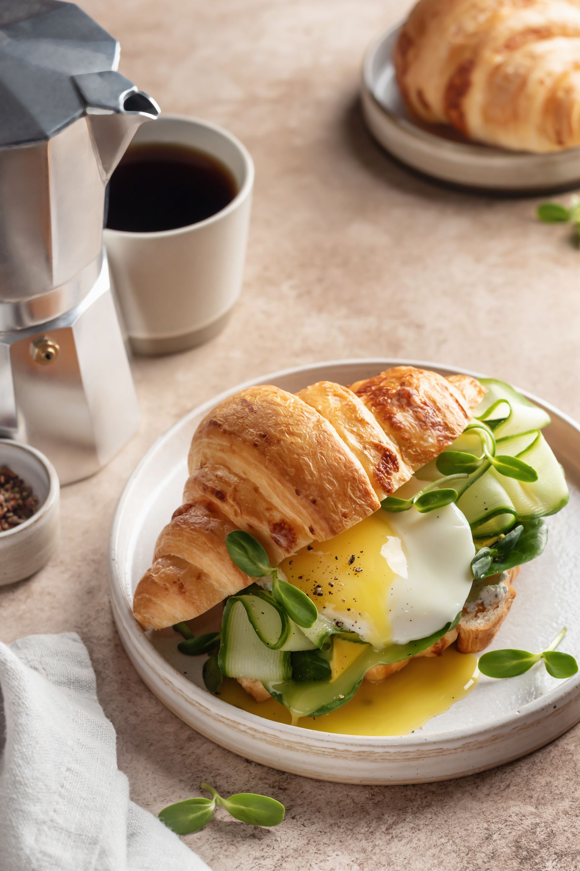 Croissant breakfast sandwich with egg, avocado, and greens on a plate beside a moka pot of coffee