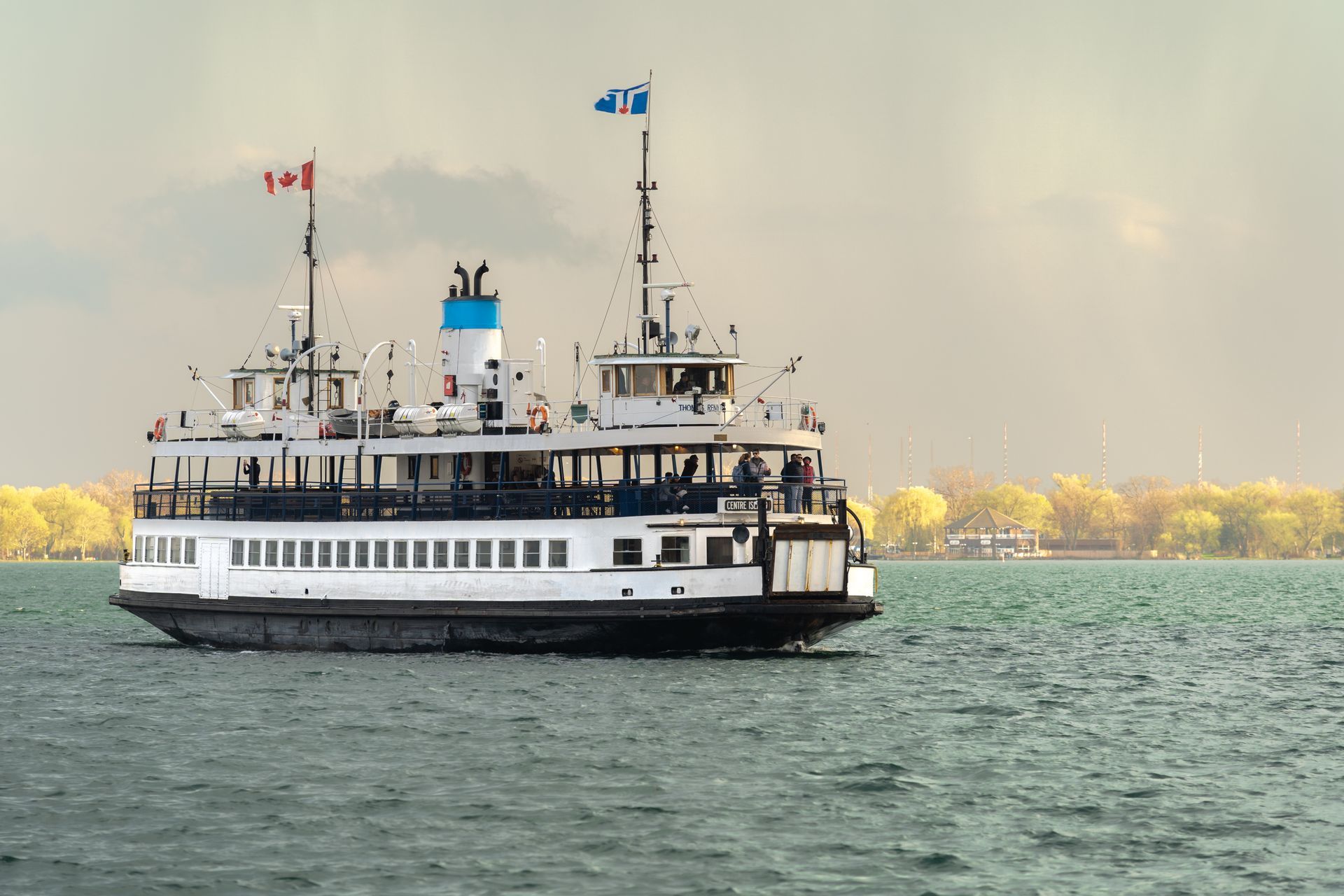 White passenger ferry on choppy water with flags, seen from the side against a distant shoreline.
