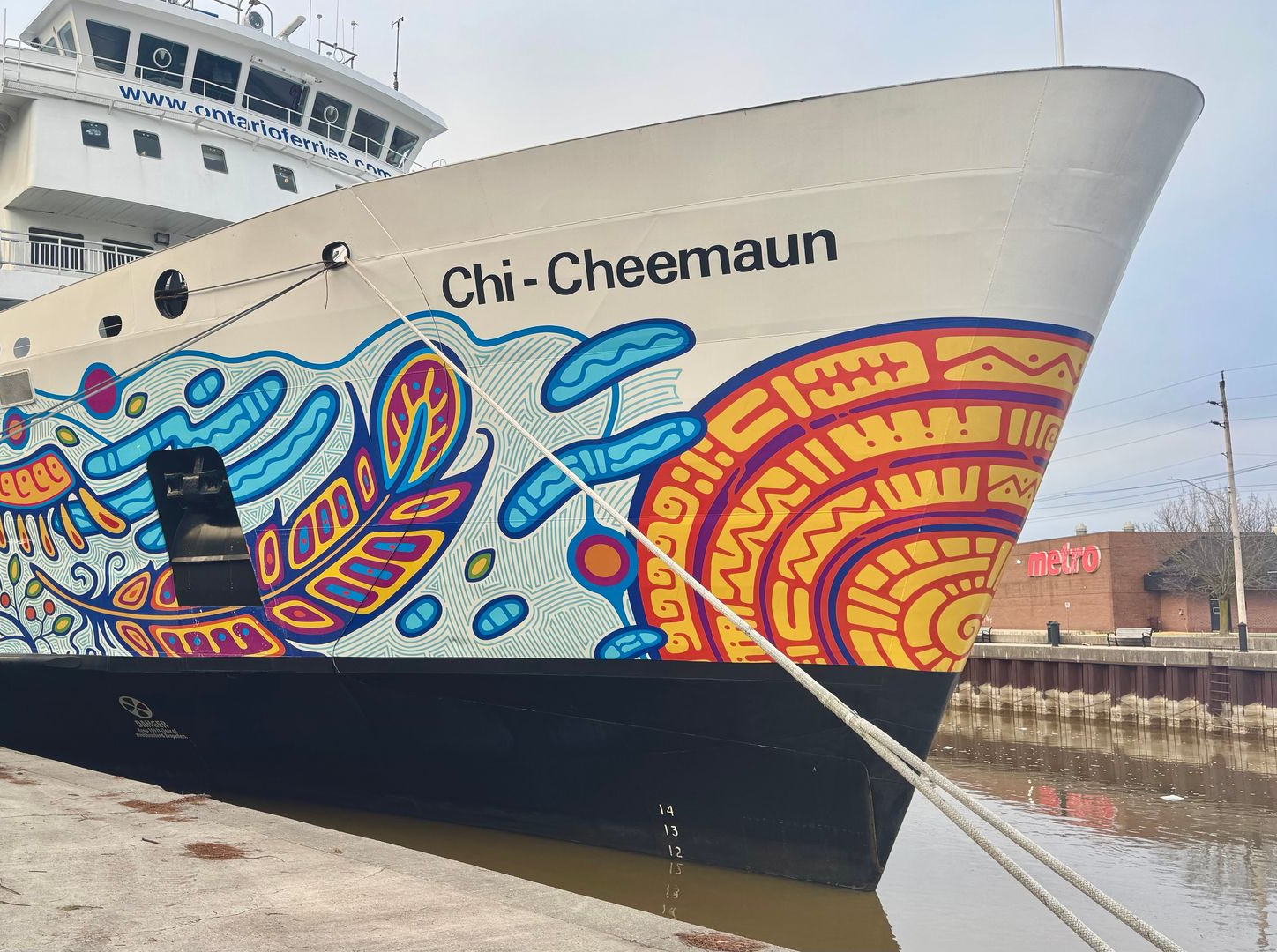 Bow of the ship Chi-Cheemaun with colorful Indigenous-style artwork docked beside a harbor wall