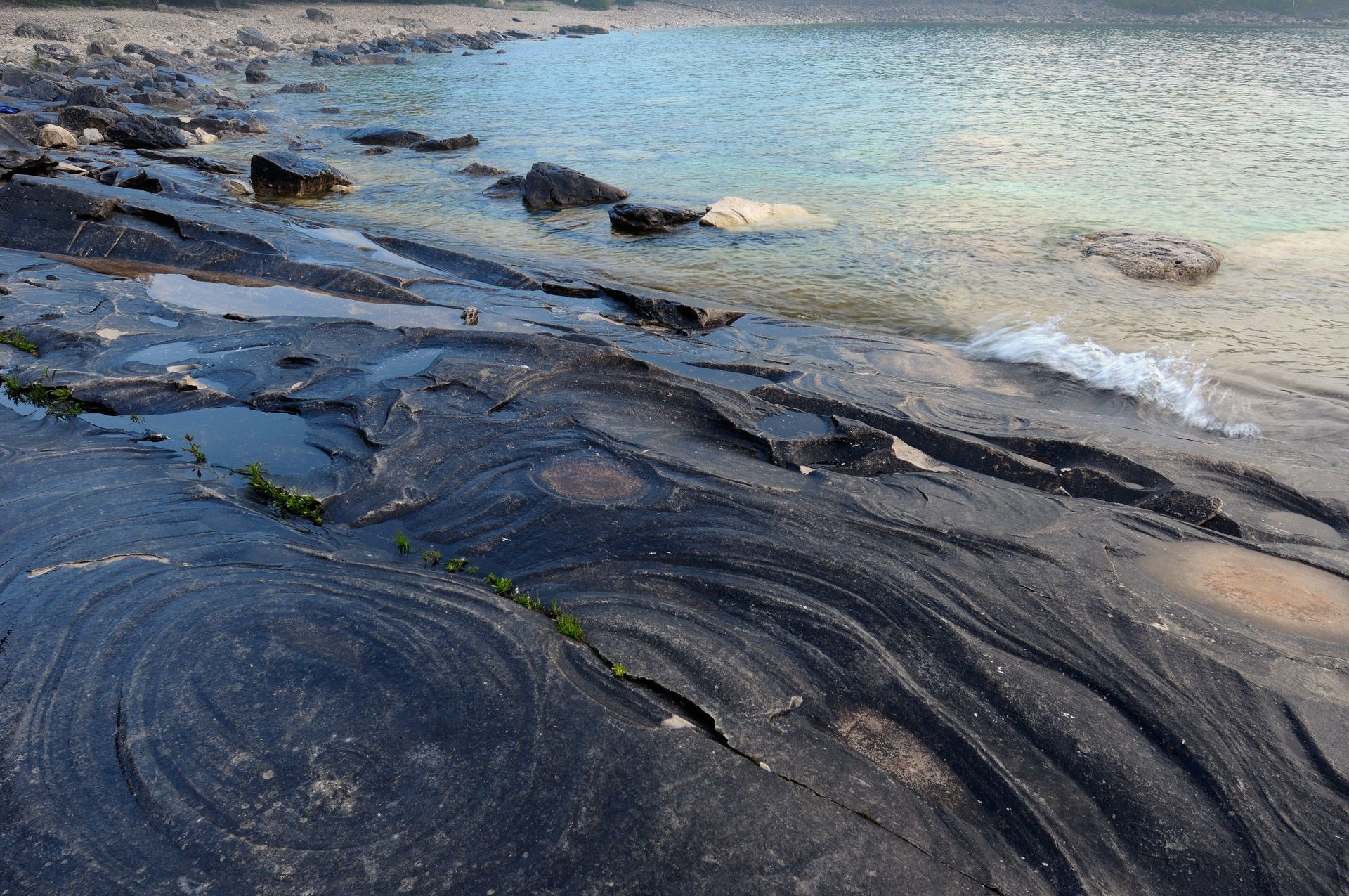 Dark layered rock ledges along a sandy shoreline with clear blue water and small waves.