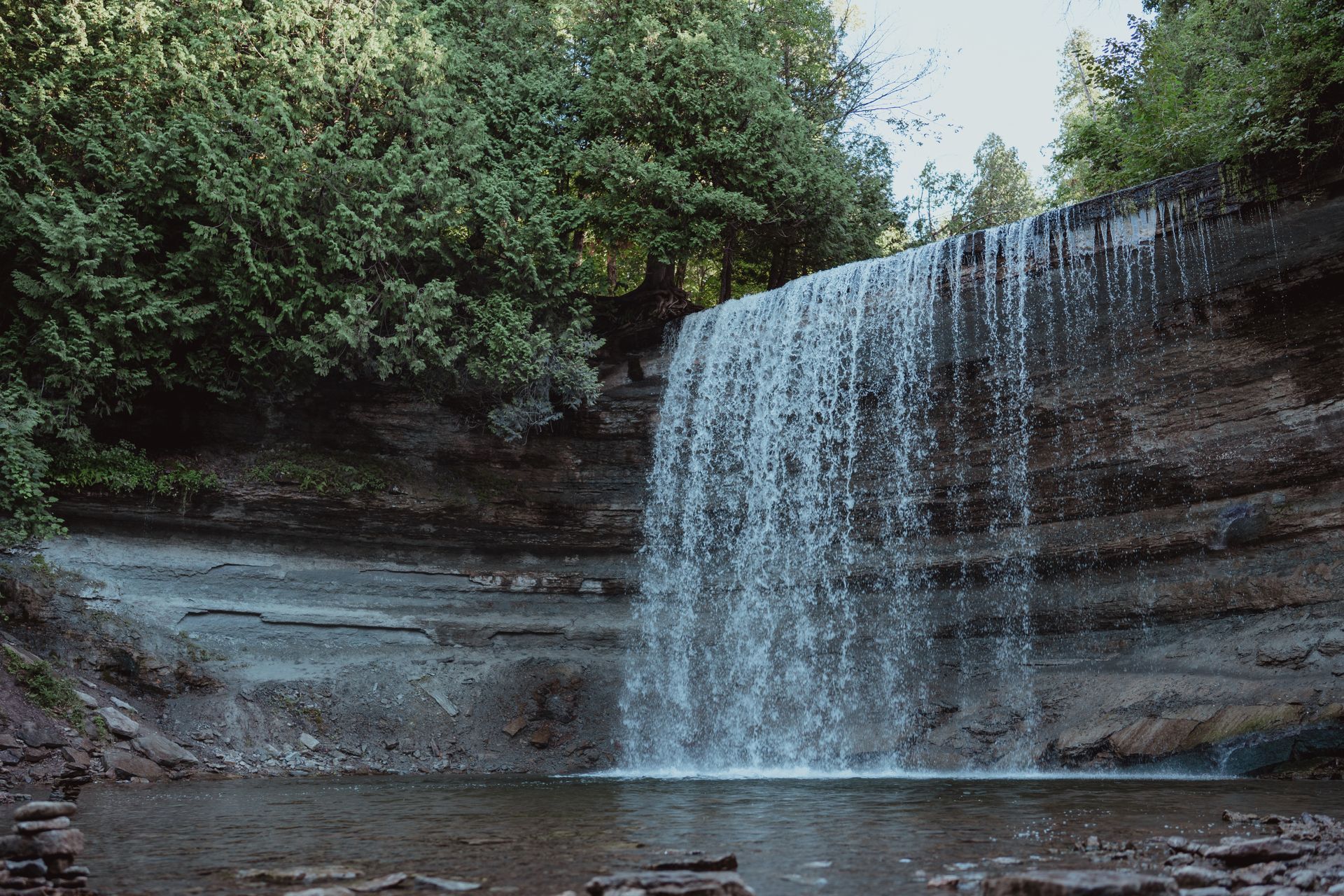 Waterfall spilling over layered rock into a small pool, surrounded by dense green forest.