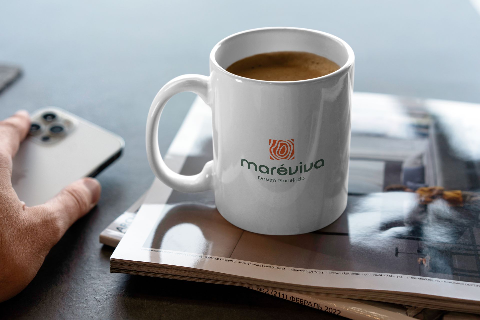 Uma caneca branca com o logotipo da Marévivo está sobre revistas ao lado de uma mão que segura um smartphone.