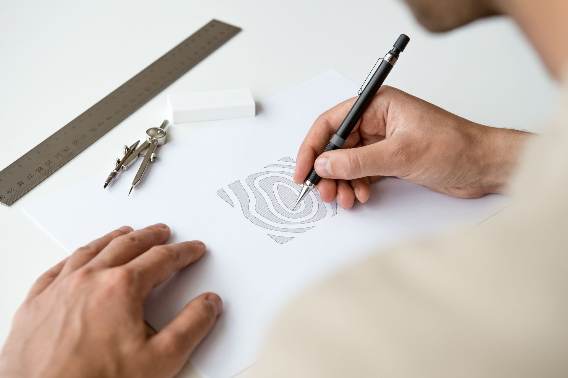 Uma pessoa usa uma lapiseira para desenhar um padrão intrincado com linhas onduladas em papel branco, com ferramentas de desenho por perto.