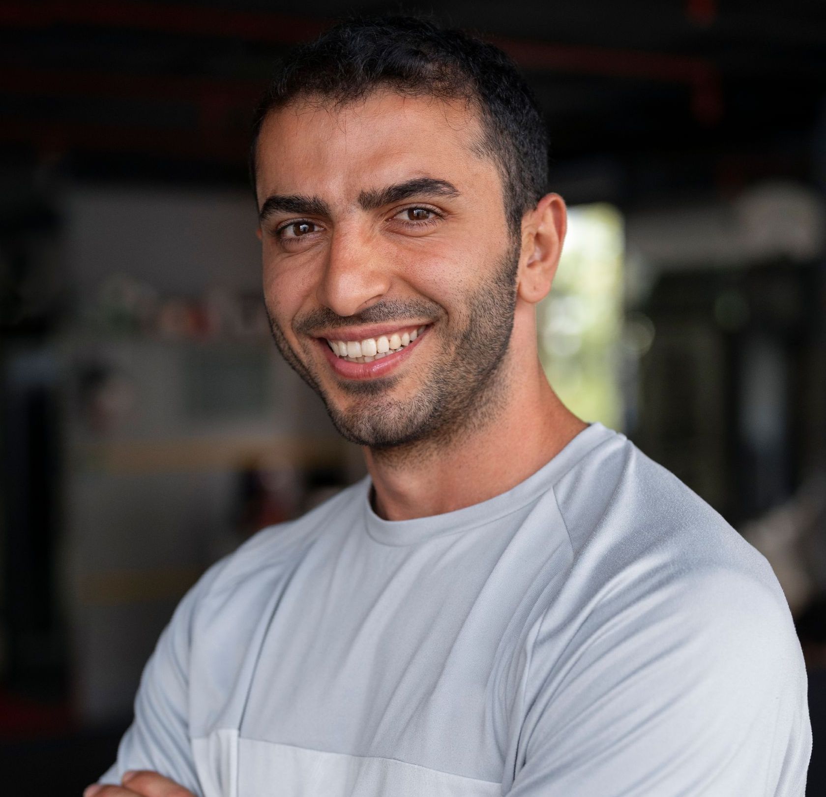 Uma pessoa sorridente vestindo uma camiseta esportiva de mangas compridas cinza-claro, fotografada do peito para cima em uma academia.