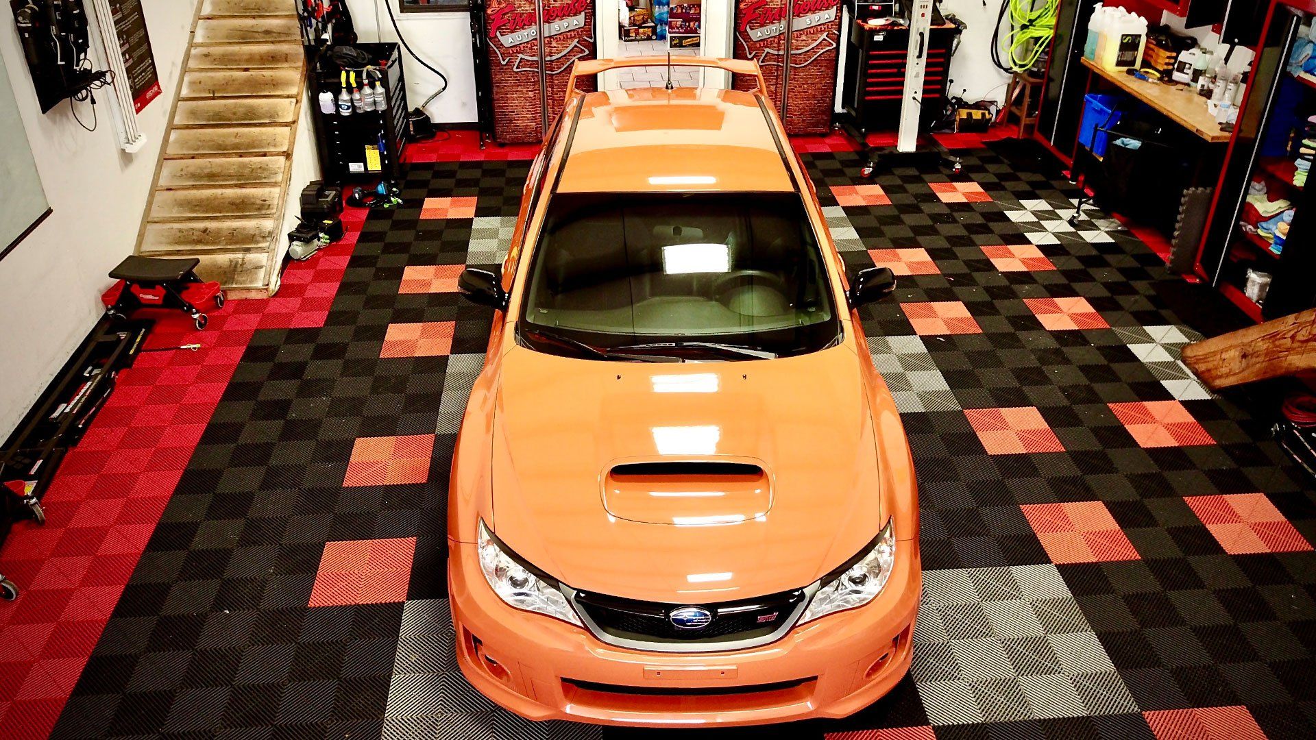 Orange car in a garage with a checkered black-and-red floor, viewed from above.