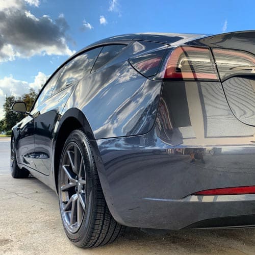 Dark gray Tesla Model 3 parked on a driveway, shown from the rear side under a cloudy sky