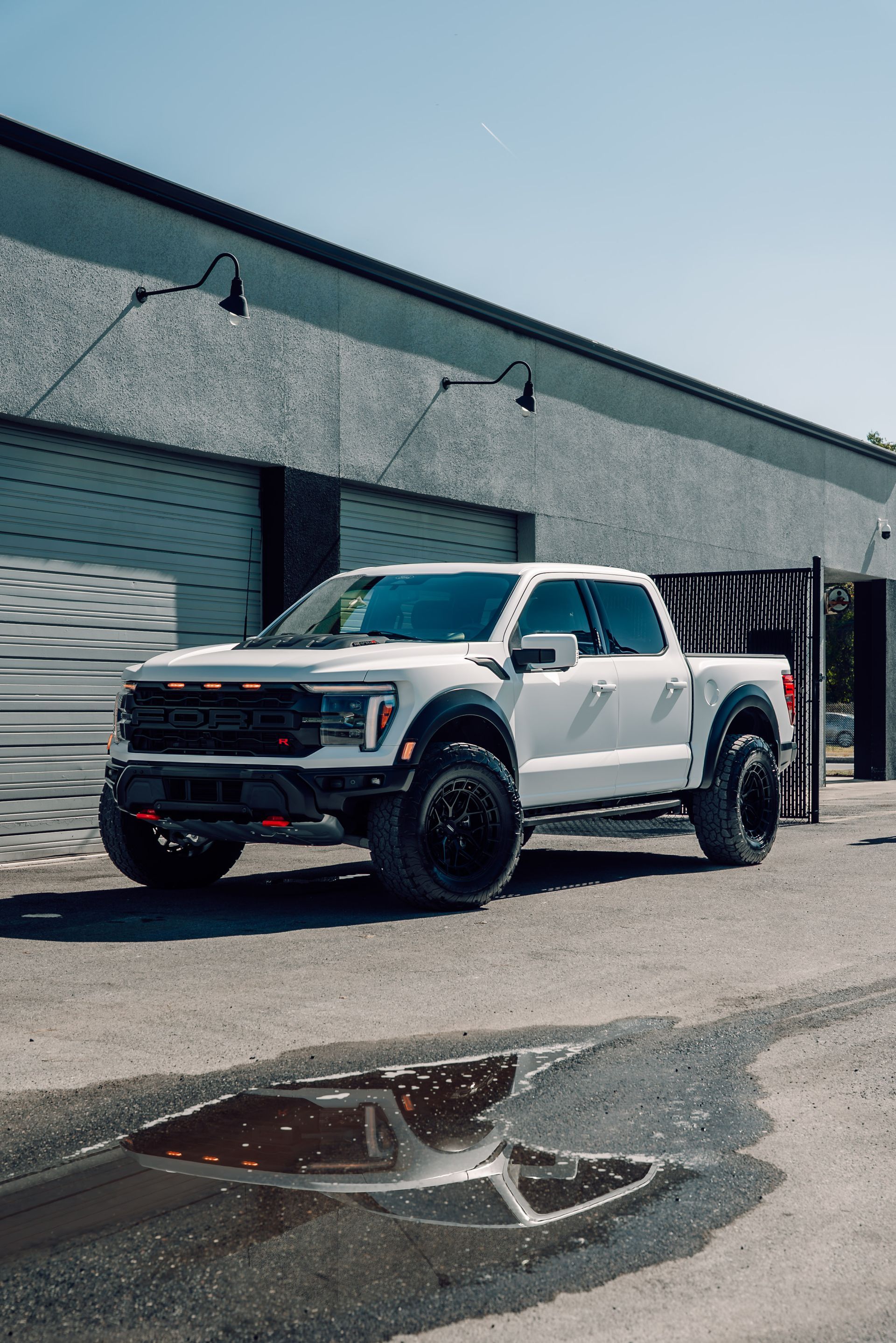 White Ford Raptor pickup truck parked in front of a gray building on a sunny day.