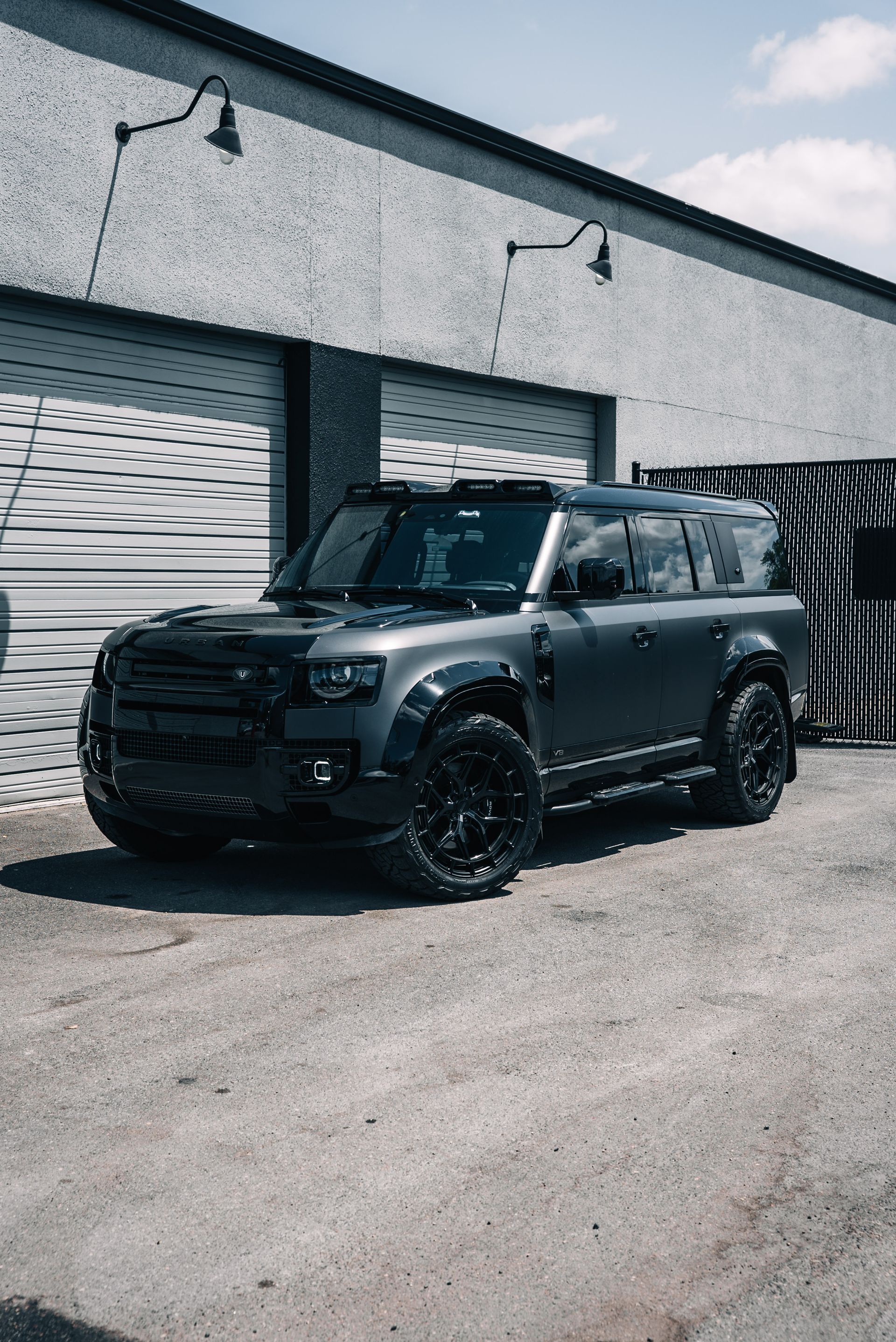 Black modified SUV parked outside a warehouse with white garage doors and a partly cloudy sky