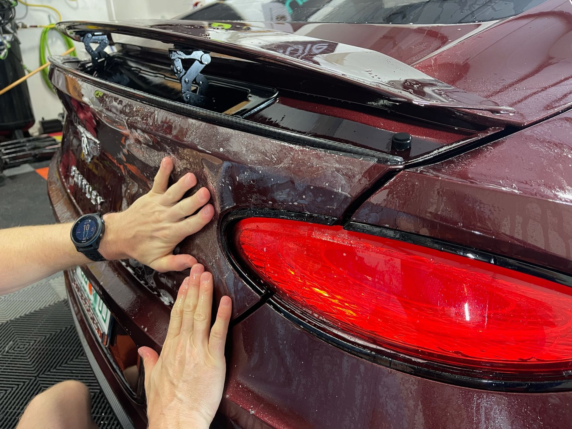 Hands pressing a maroon car trunk panel beside a red taillight in a workshop