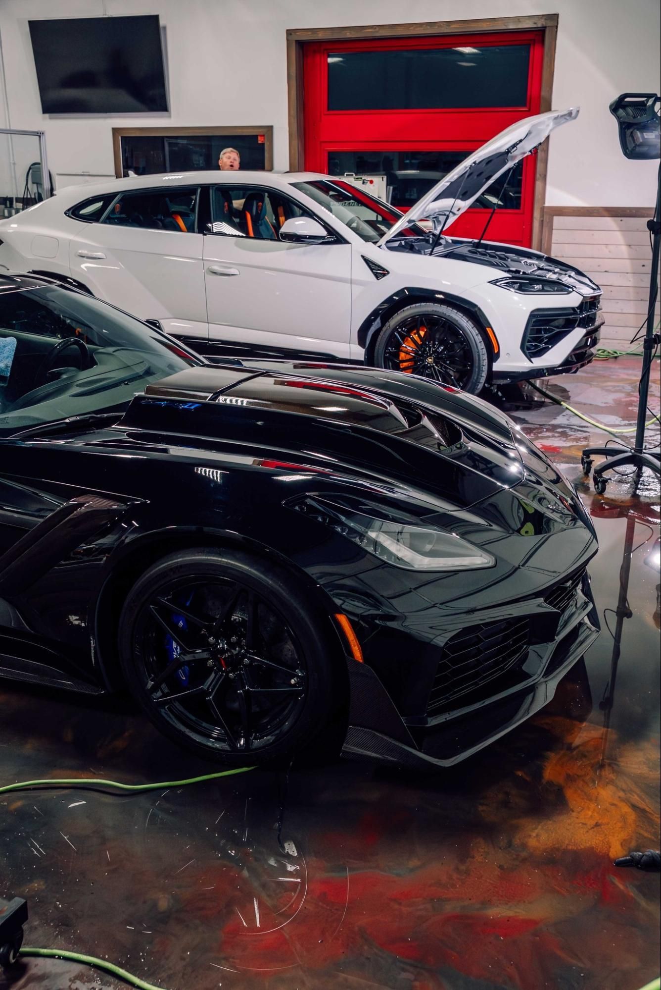 Black sports car in foreground and white SUV in garage, both with hoods open for repair or tuning.