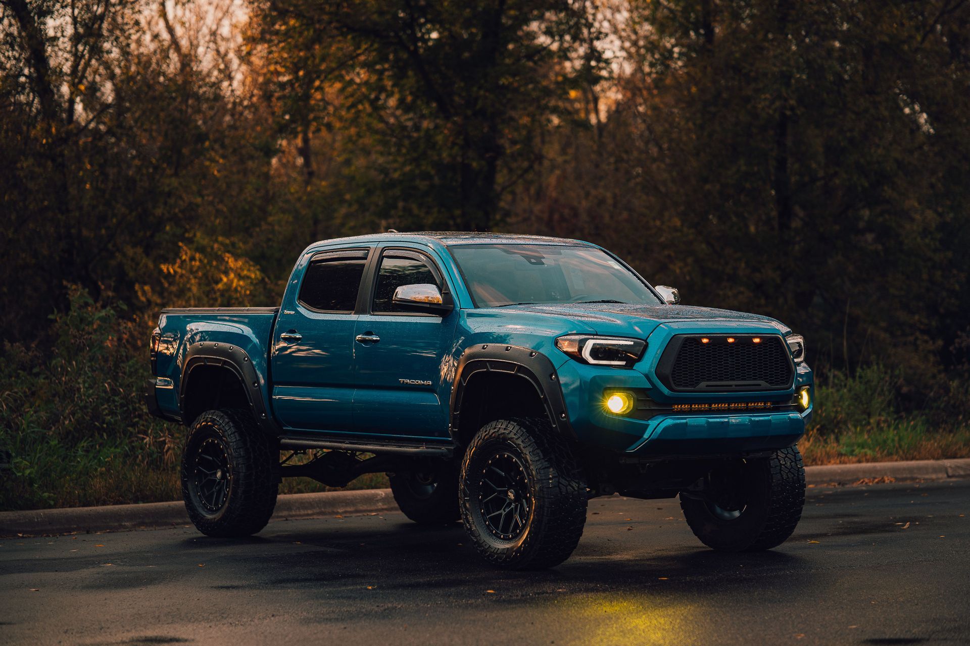 Blue lifted pickup truck parked on a wet road at dusk in a wooded area