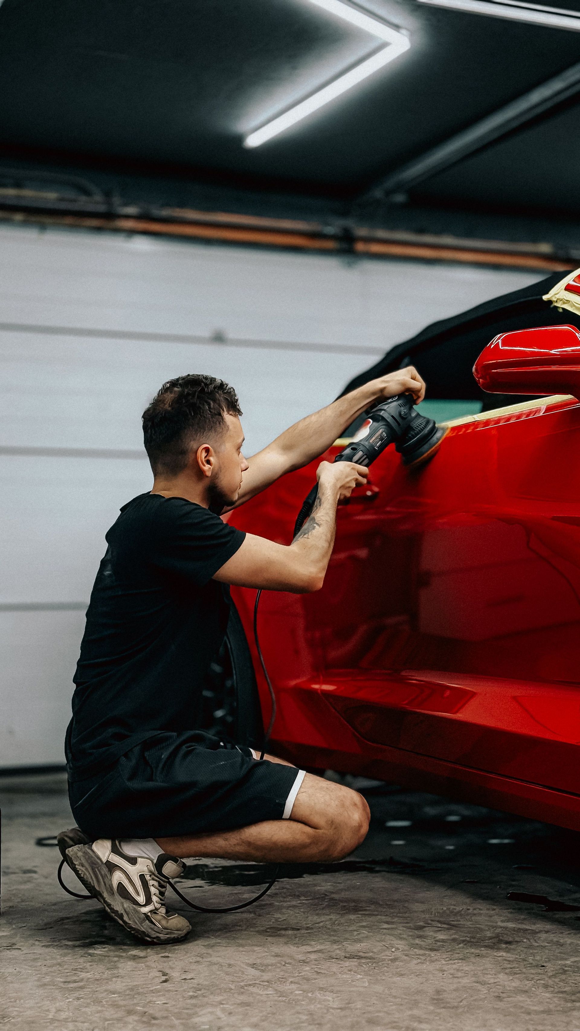 Person kneeling beside a red sports car, polishing the rear quarter panel in a garage