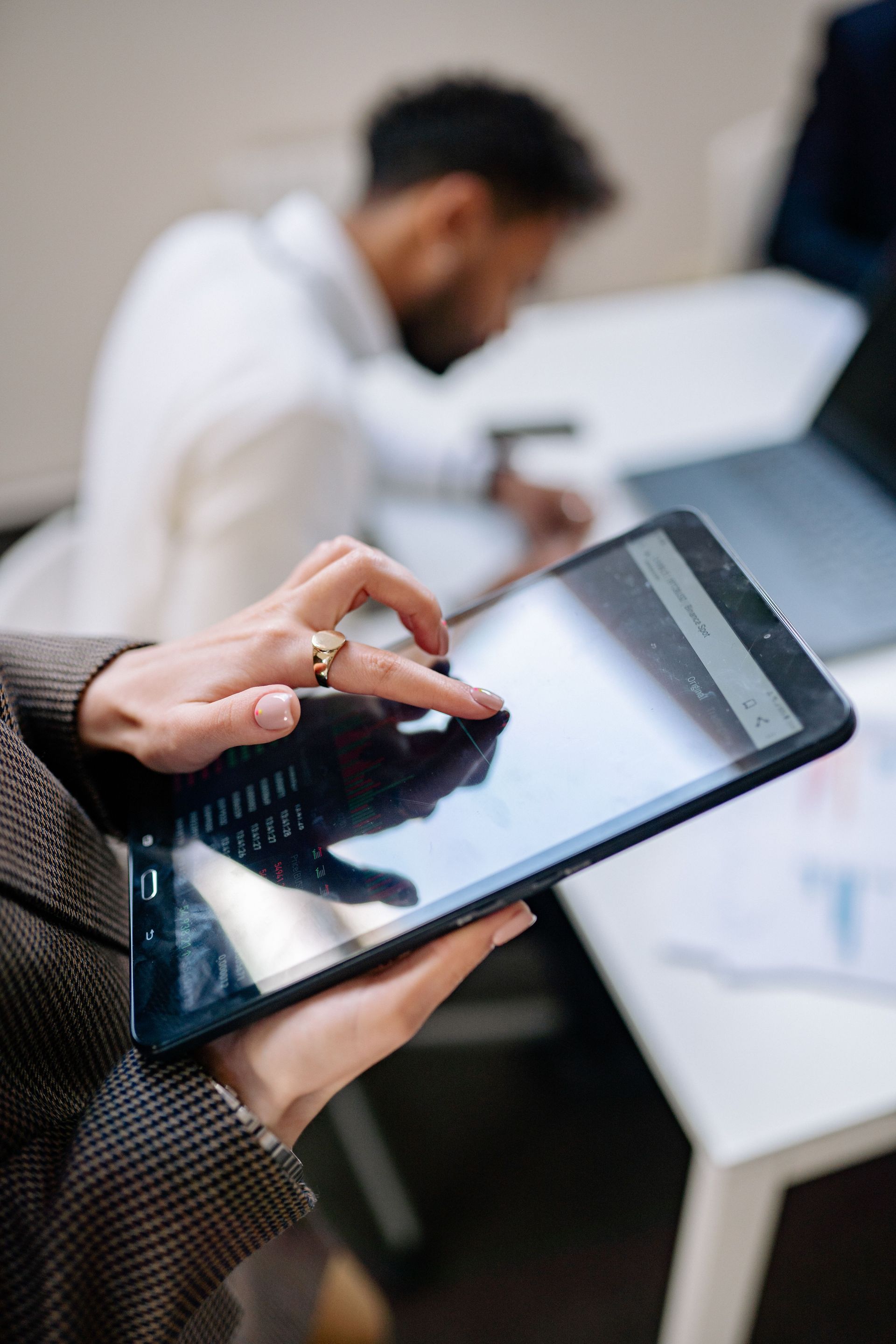 Person using a tablet with a stylus at a desk, with a blurred coworker and laptop in the background