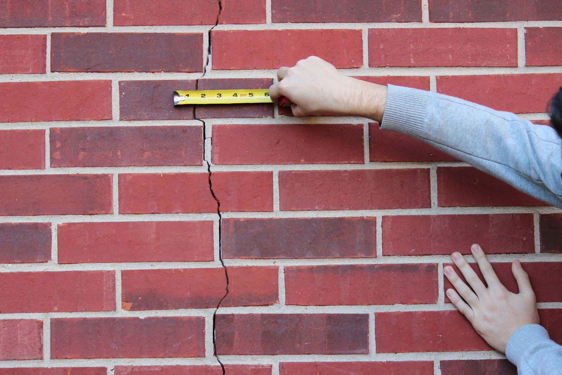 Hands measuring a cracked brick wall with a yellow level and ruler