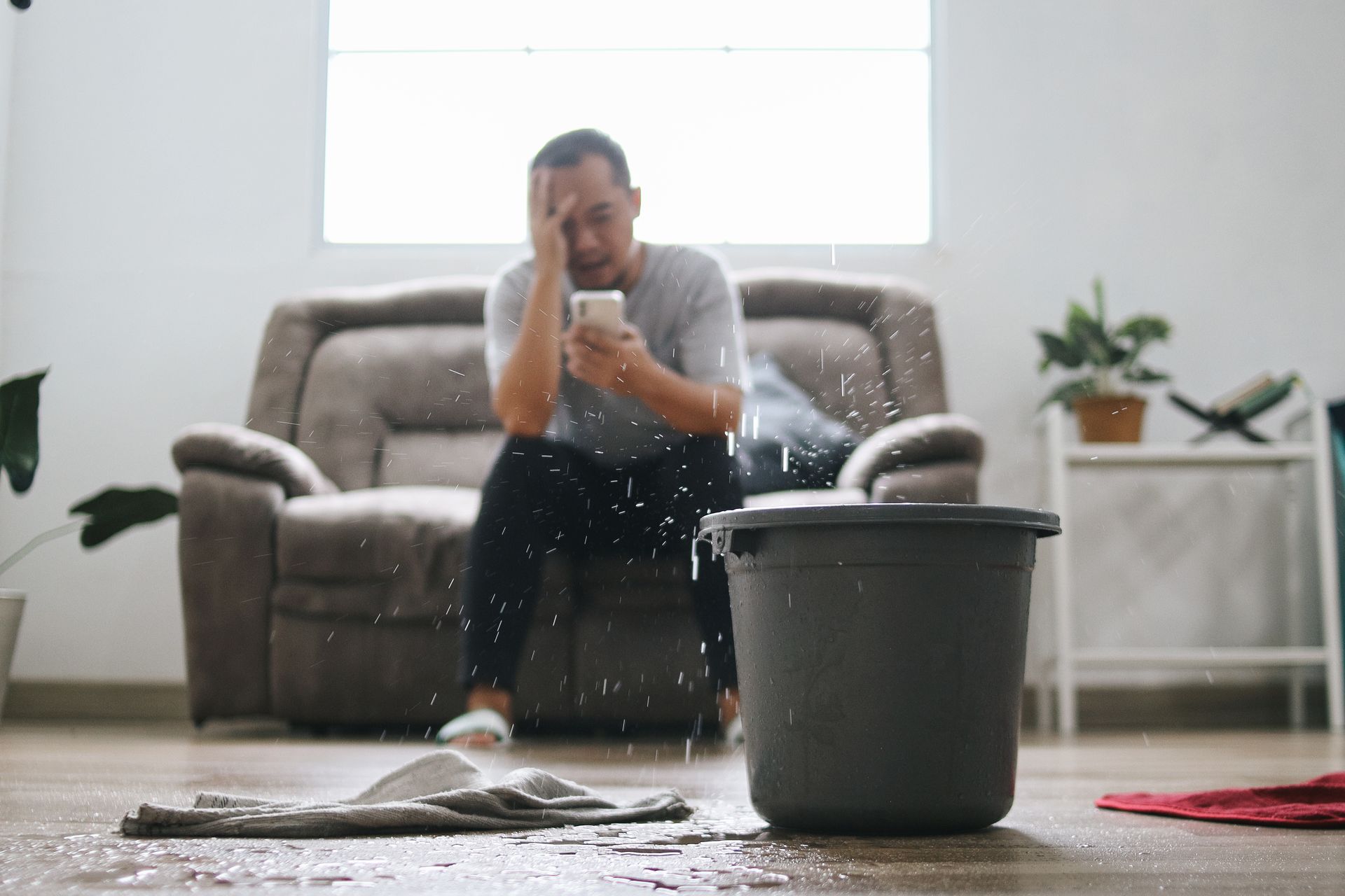 Person sitting on a living room floor with a bucket, cleaning up spilled water near a couch.