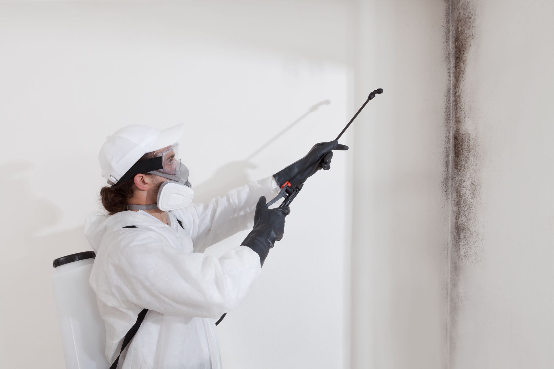 Worker in white protective gear using a sprayer on a wall corner