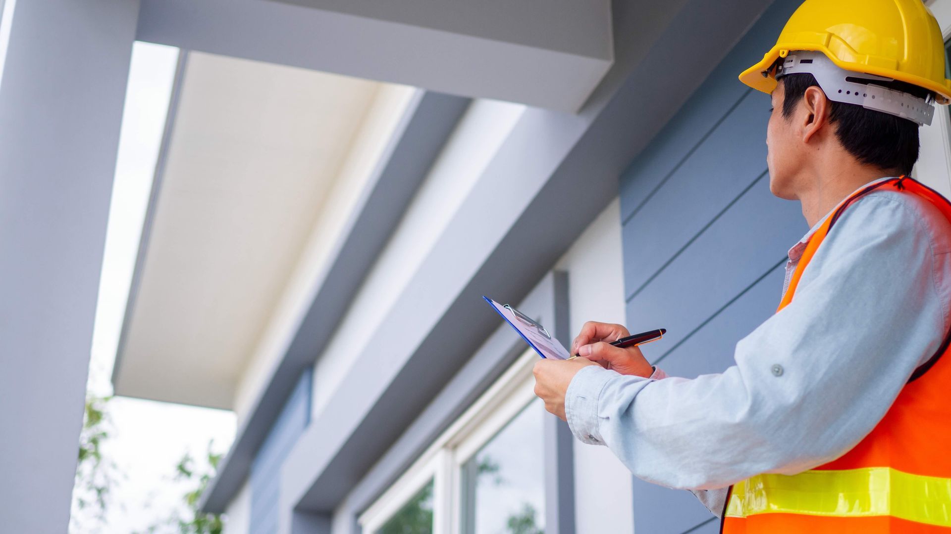 a man wearing a hard hat and safety vest is writing on a clipboard in front of a building .