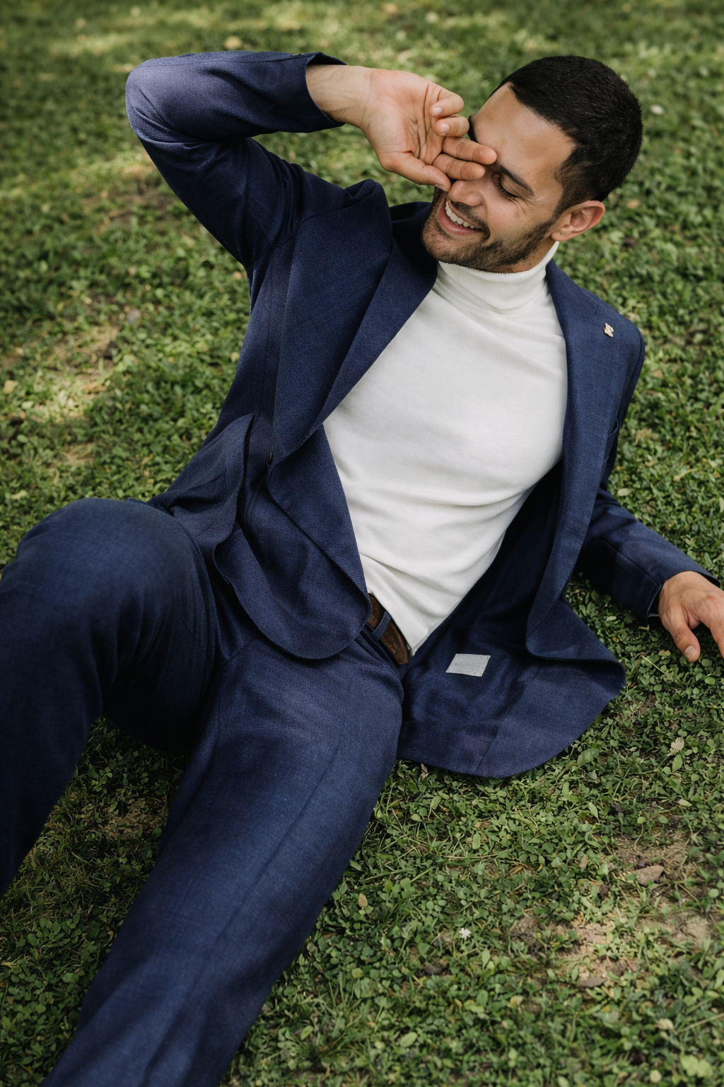Un uomo che indossa un abito blu e un dolcevita bianco sorride, sdraiato sull'erba e si ripara gli occhi.