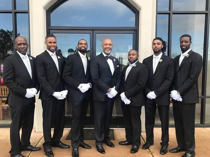 Seven men in black tuxedos and white gloves standing in front of a building entrance.