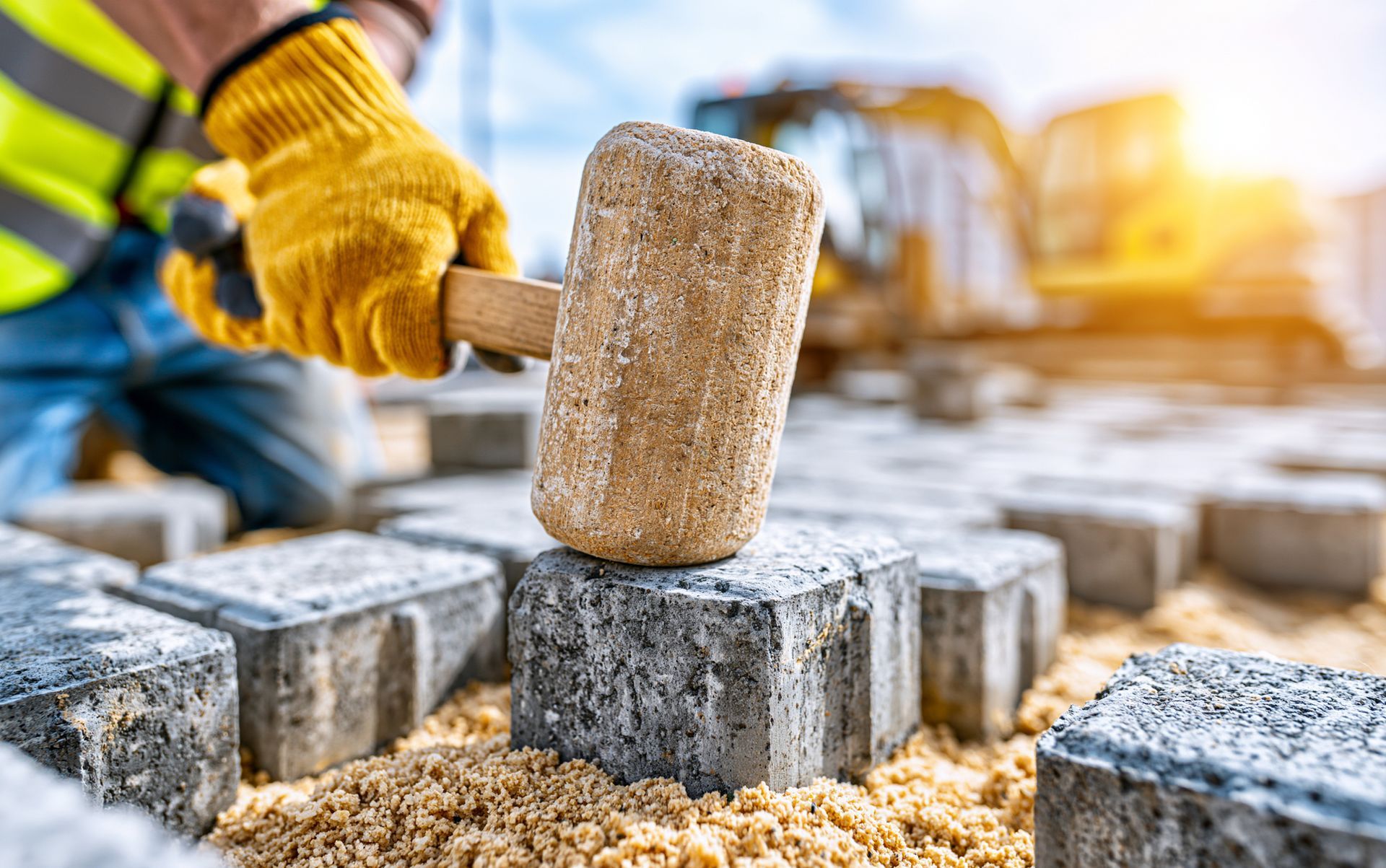 Worker setting a gray paver with a rubber mallet on a sand base.