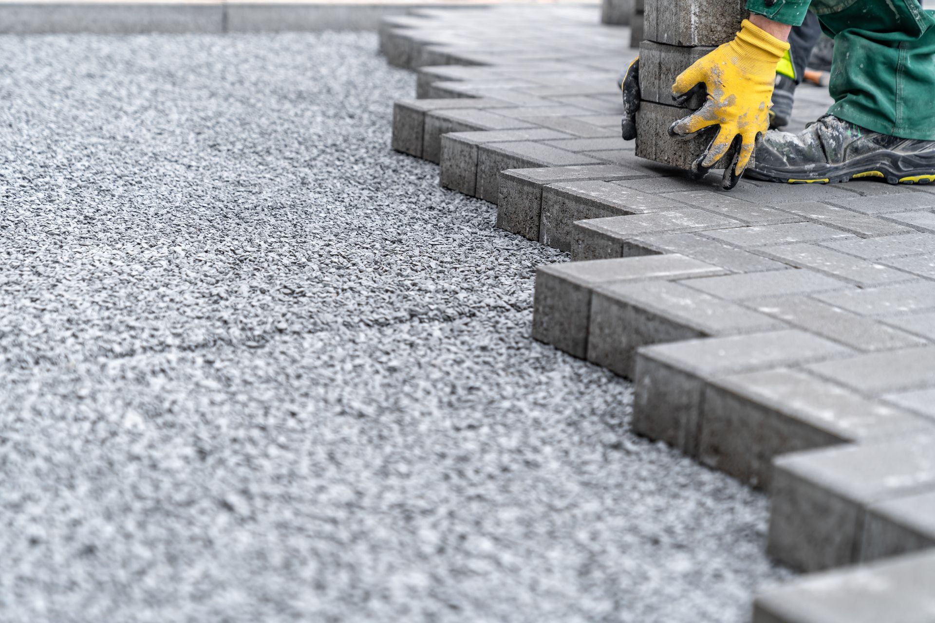 Worker laying gray concrete paving stones on gravel base, professional brick patio installation.