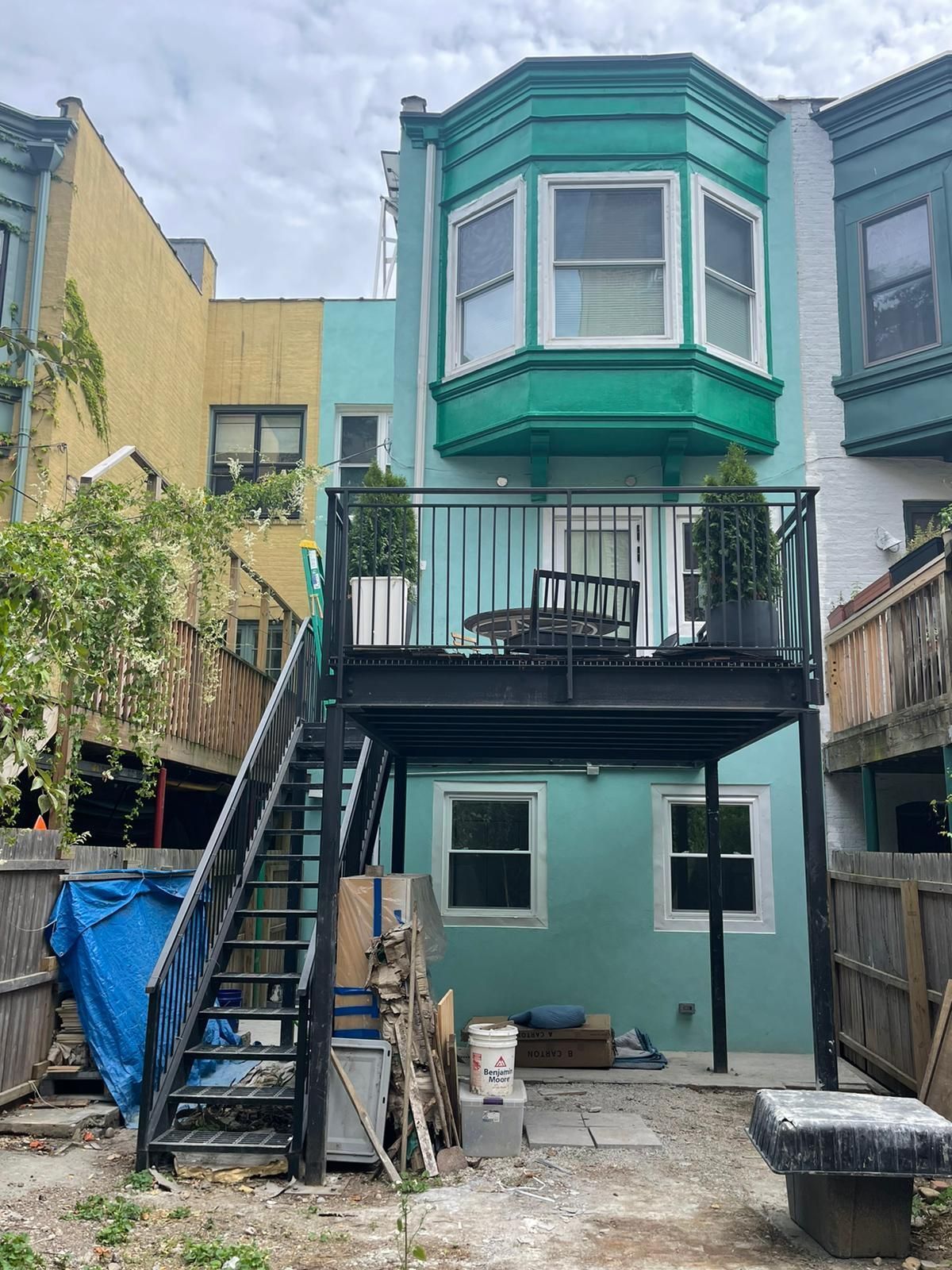 Narrow backyard with a teal two-story house, exterior stairs, and a small deck above a lower patio.