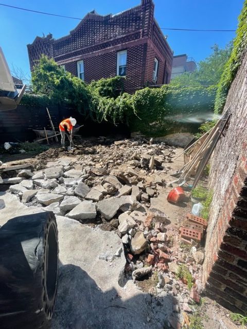 Worker clearing rubble in a narrow brick-walled yard beside a house.