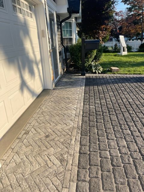 Brick-paved driveway beside a white garage and house, leading to a lawn with a white bench and trees.