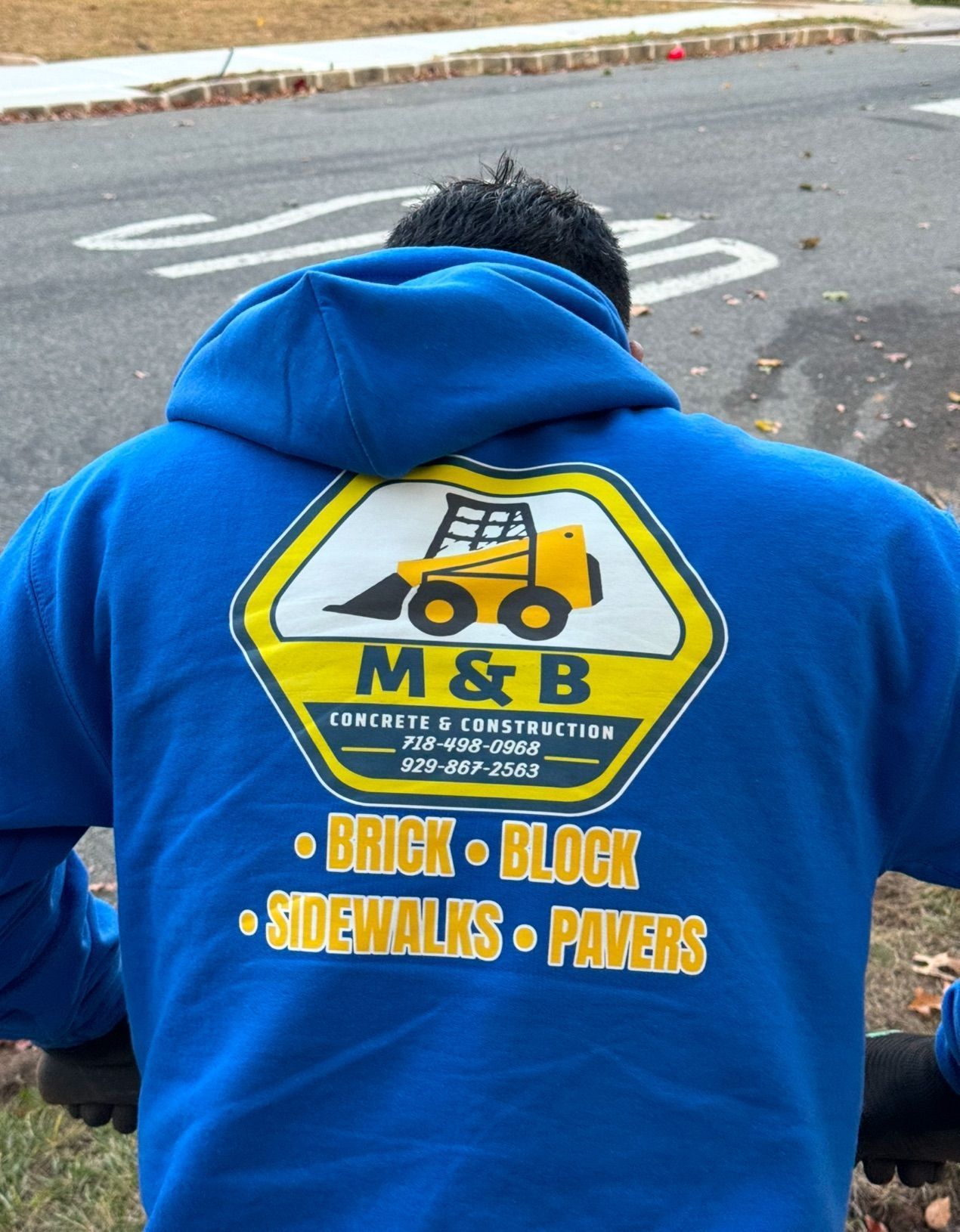 Person in blue hoodie with M&B brick and block paving logo on the back, sitting outdoors on pavement.