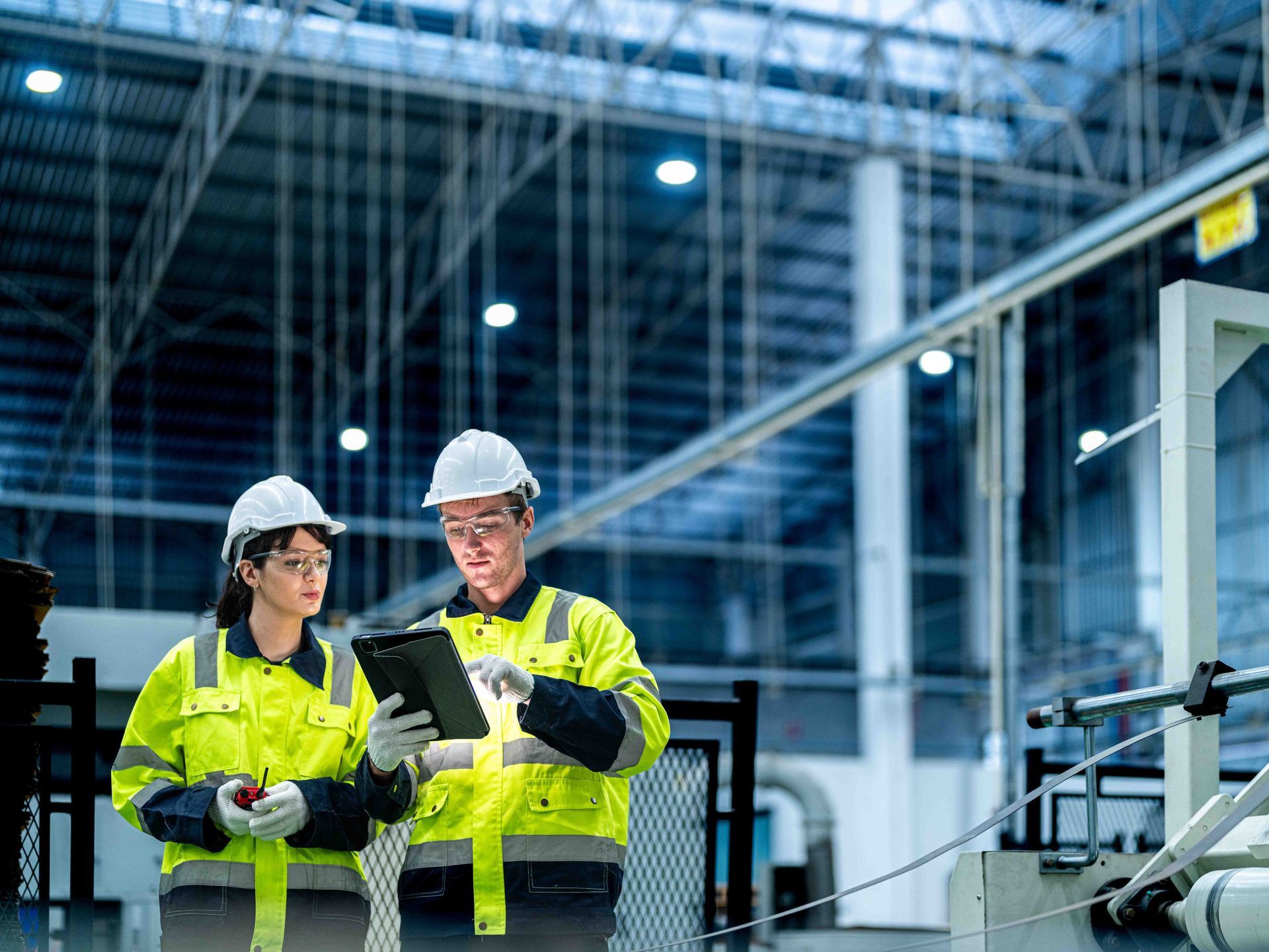 Two workers in hard hats and safety vests reviewing a tablet in a factory.