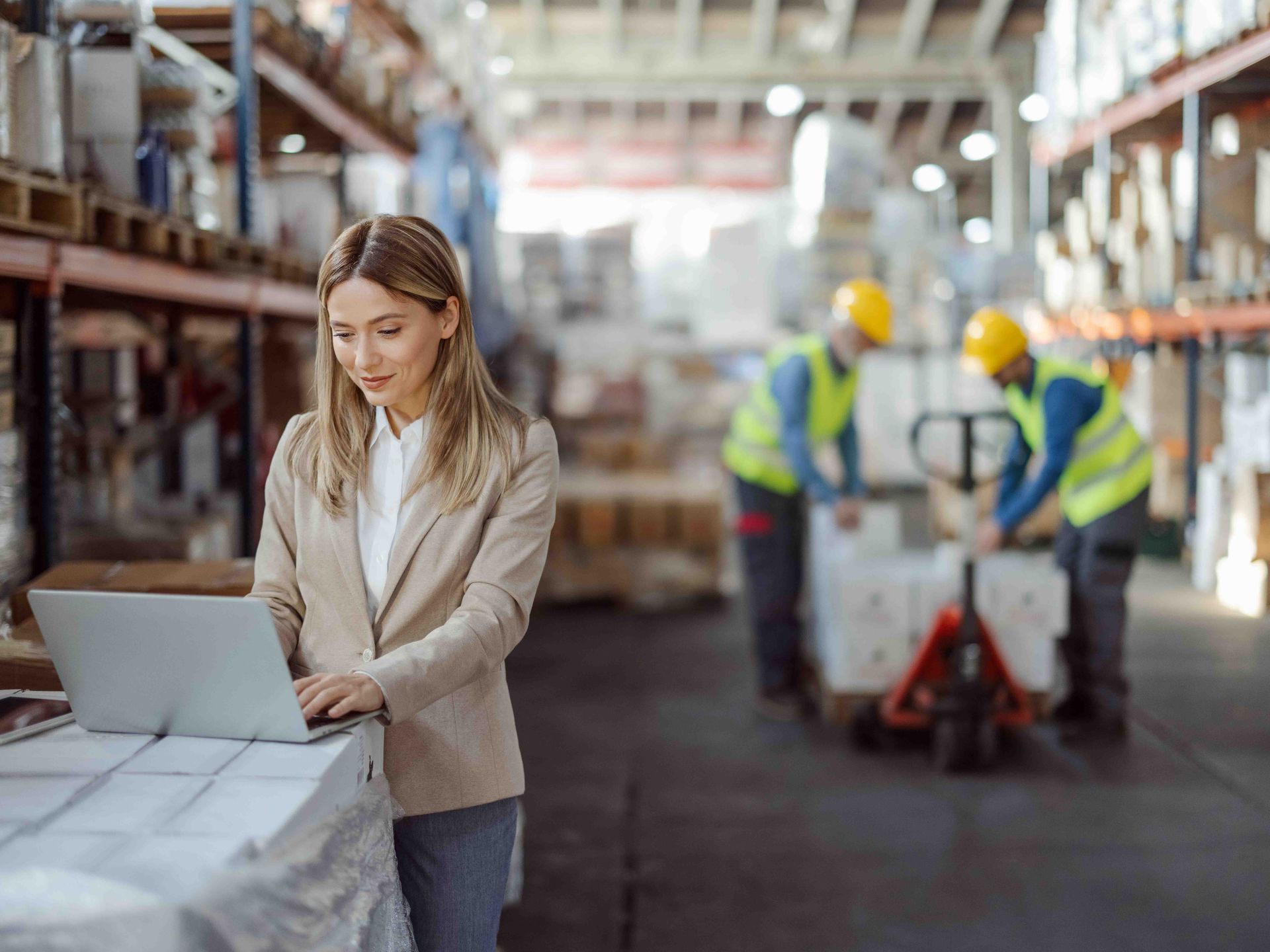 Woman in blazer using laptop in warehouse, workers loading pallets.
