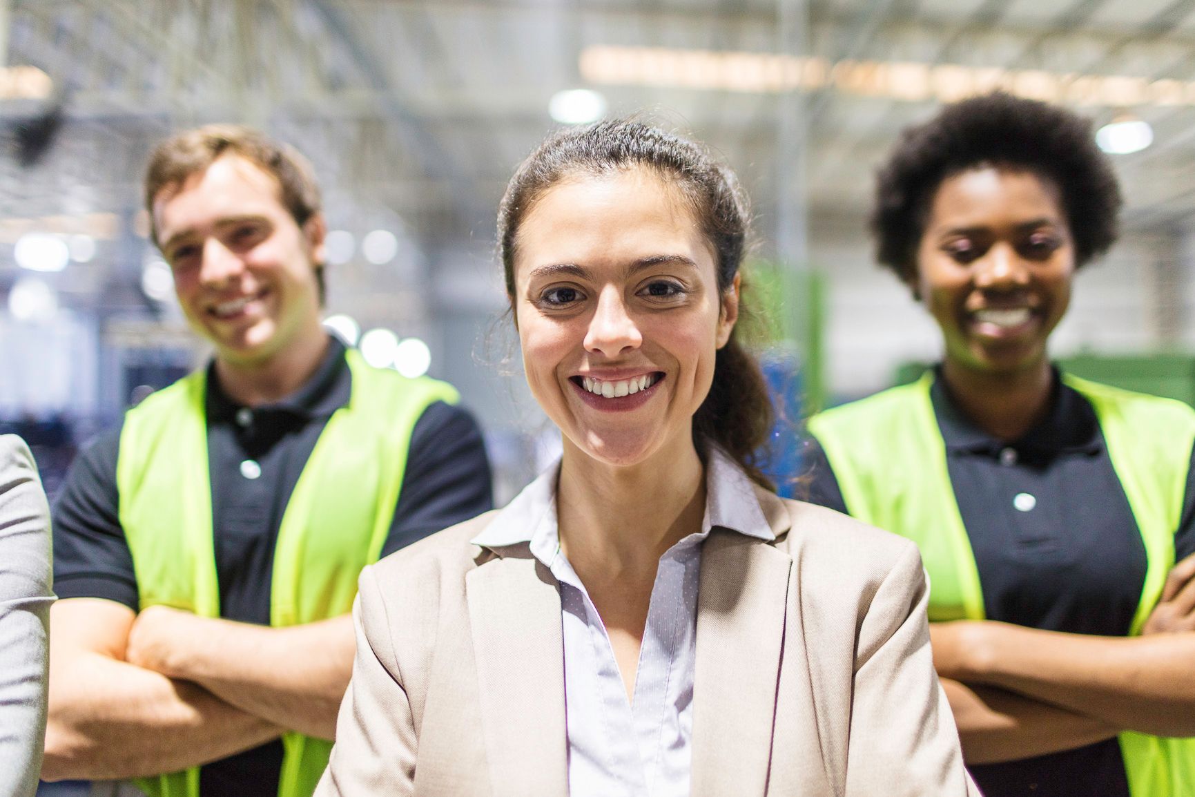 Three smiling people, arms crossed, in a warehouse setting. One in a blazer, two in safety vests.