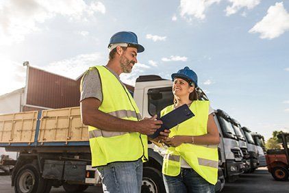 Two people in safety vests and hard hats reviewing paperwork in a truck yard.