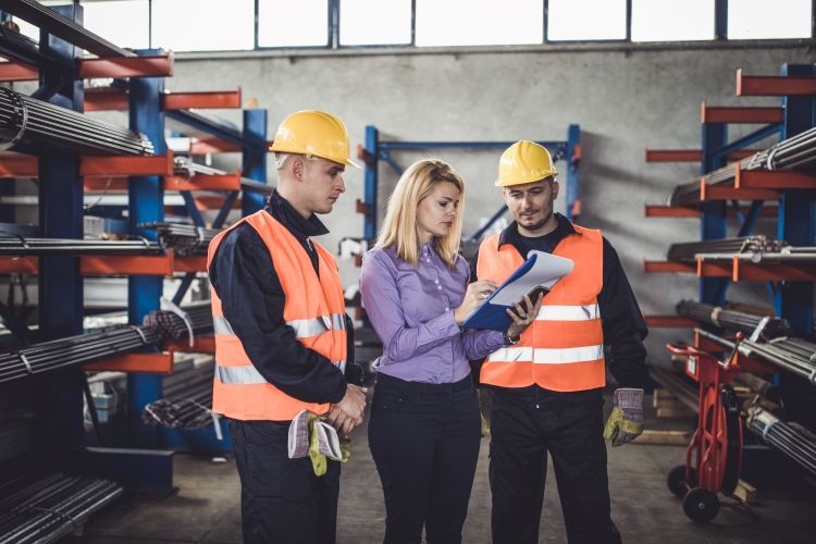 Woman in purple shirt reviews documents with two workers wearing safety vests and hard hats in a warehouse.